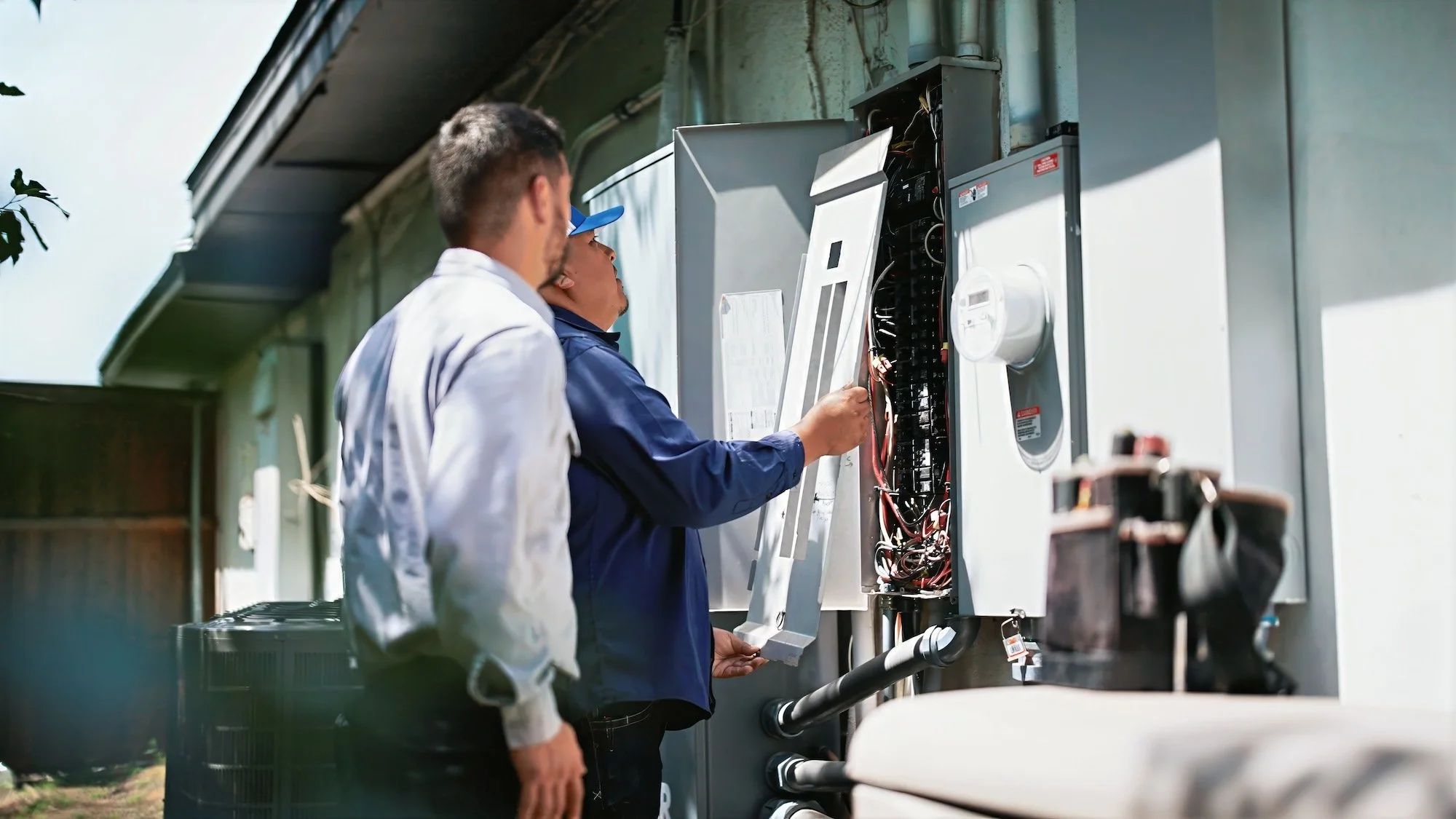 Two electricians working on an outdoor electrical panel, with one holding a tool and inspecting the wiring inside.