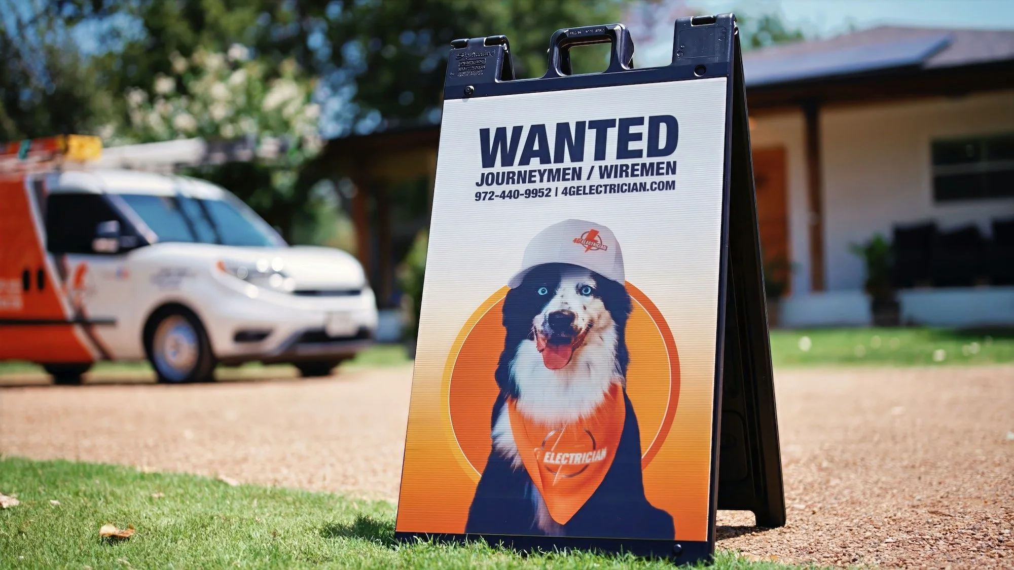 A sidewalk sign advertising a wanted electrical journeyman or wireman, with a picture of a dog wearing a construction helmet and bandana, parked in front of a house with a tree and a vehicle in the background.