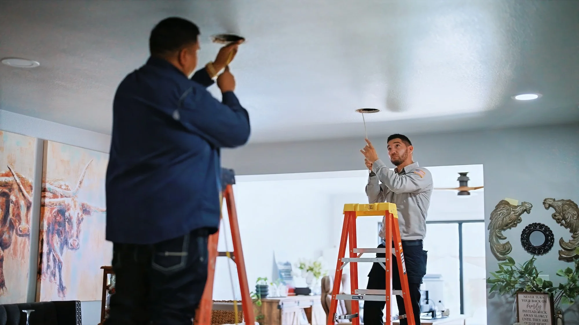 Two men installing or repairing a ceiling light fixture in a living room, standing on ladders and focusing on the task.