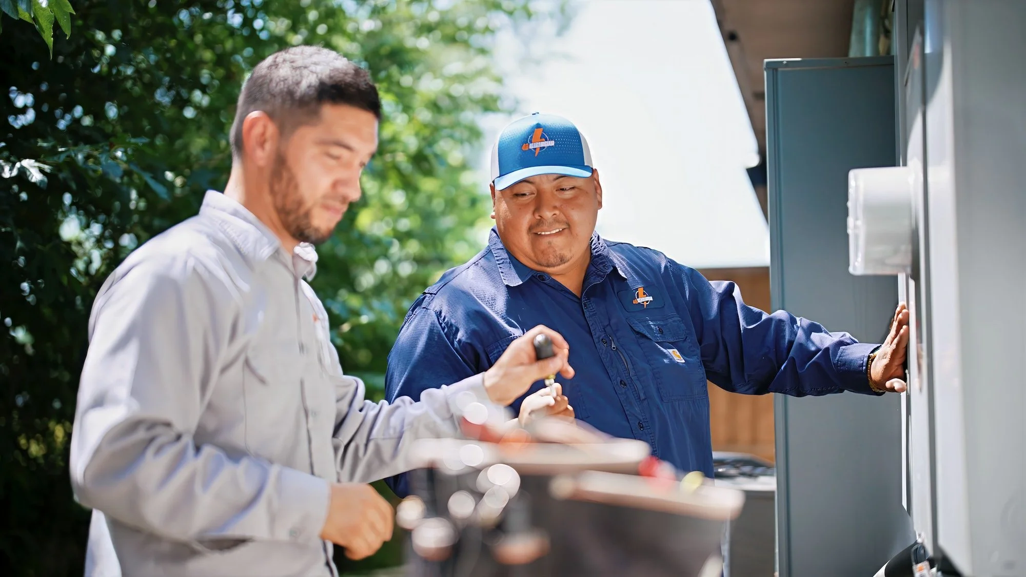 Two men, one with a beard and one clean-shaven, working together outdoors near a utility box or electrical panel. The man on the right is wearing a blue uniform and cap, and appears to be explaining something to the man on the left, who is wearing a