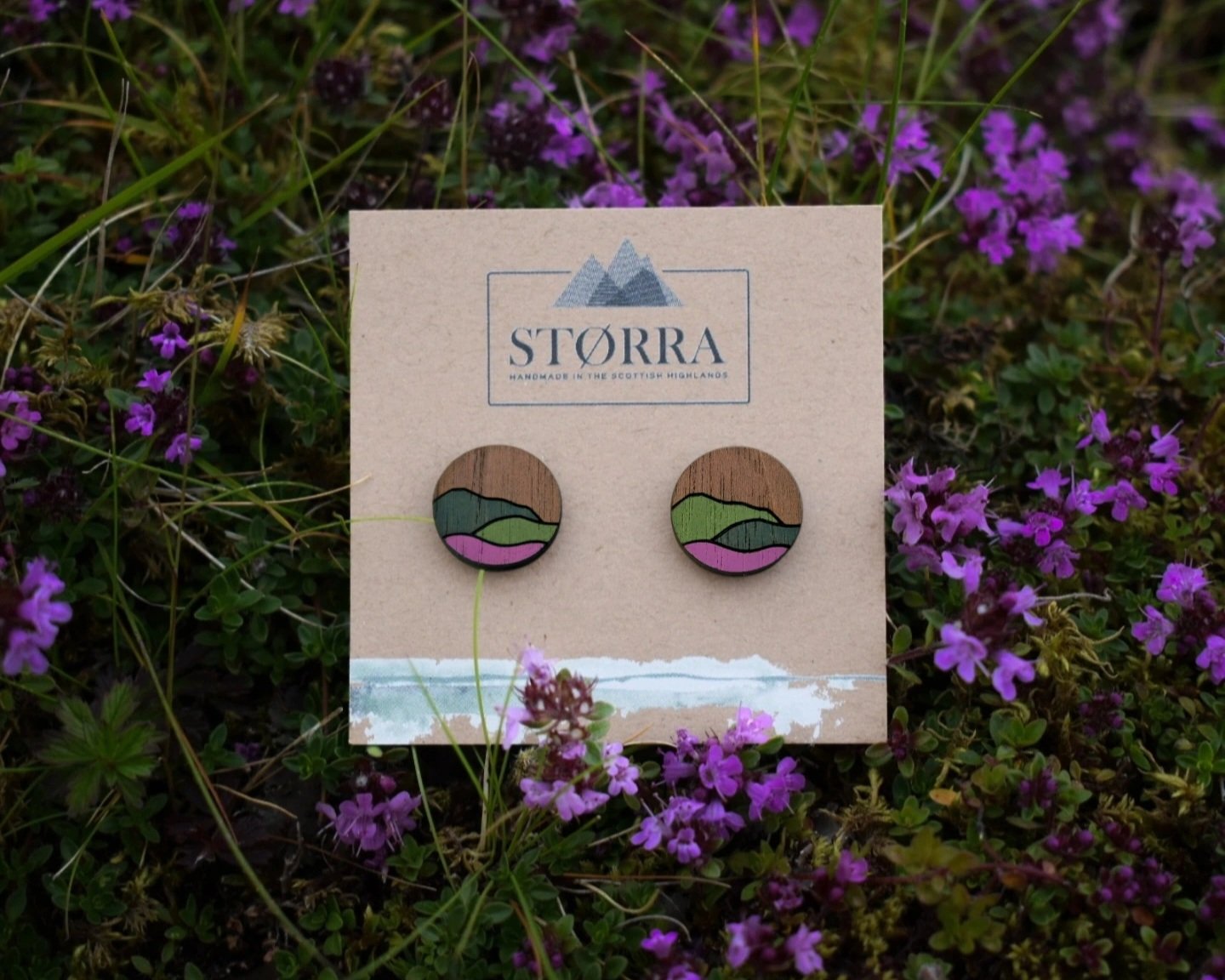 Pair of round wooden earrings with painted landscape scenes featuring mountains, green fields, and pink hills, displayed on a beige card with 'STORRA' logo, amidst purple flowers.