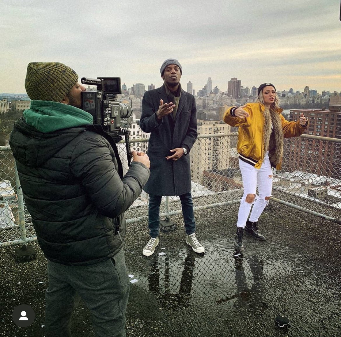 Three people on a rooftop with cityscape in the background, one person filming with a camera while the other two pose, wearing winter clothing.