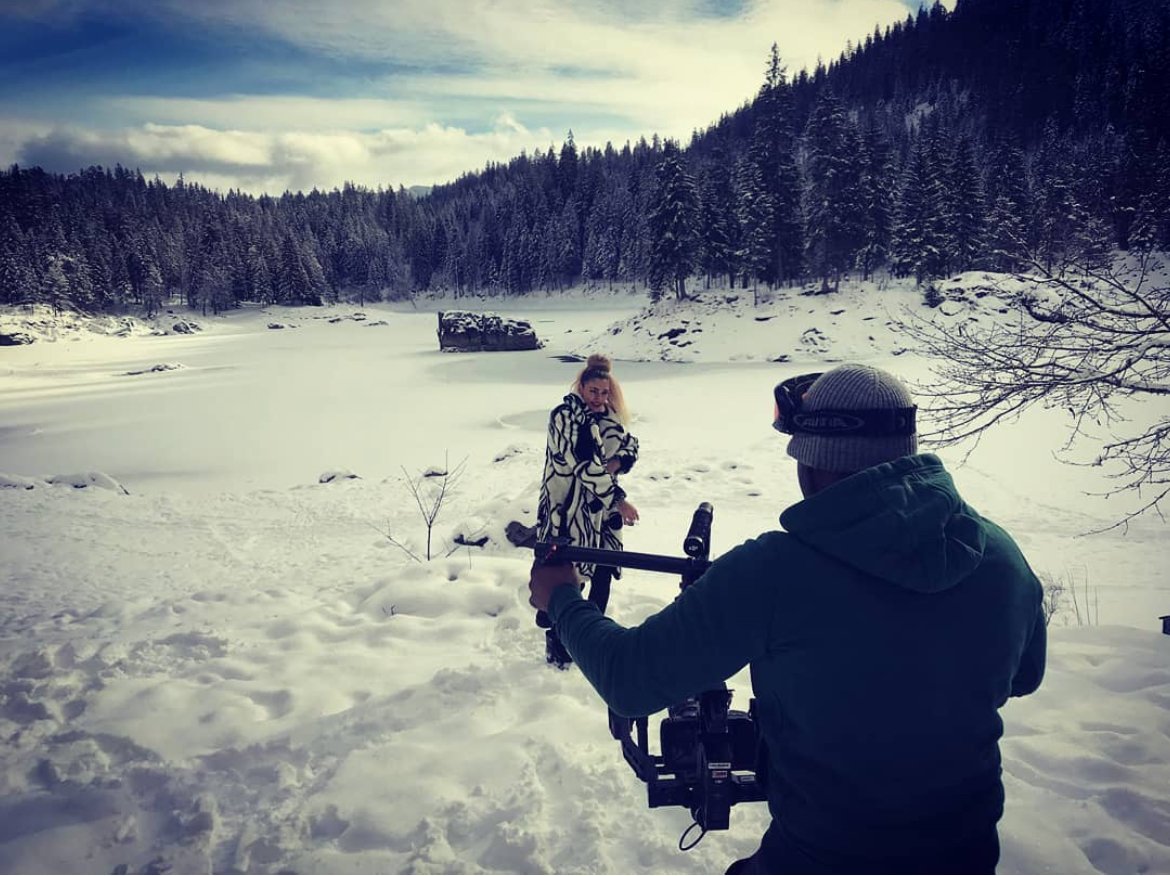 Cameraman filming a person wearing a patterned coat in a snowy forest landscape.