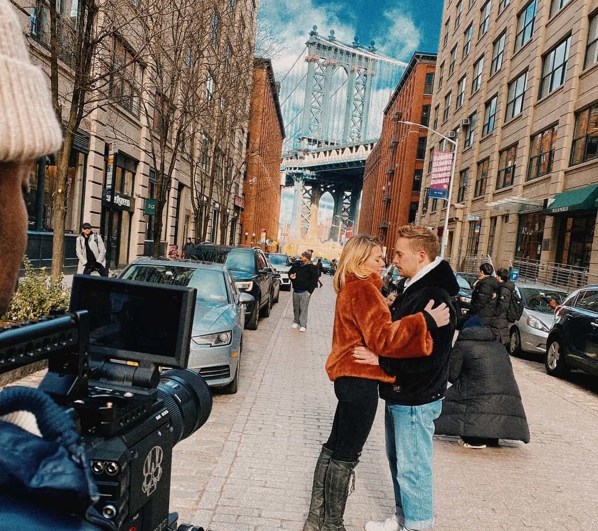 Couple posing on a street with the Manhattan Bridge in the background, surrounded by parked cars and urban buildings, with a camera visible in the foreground.