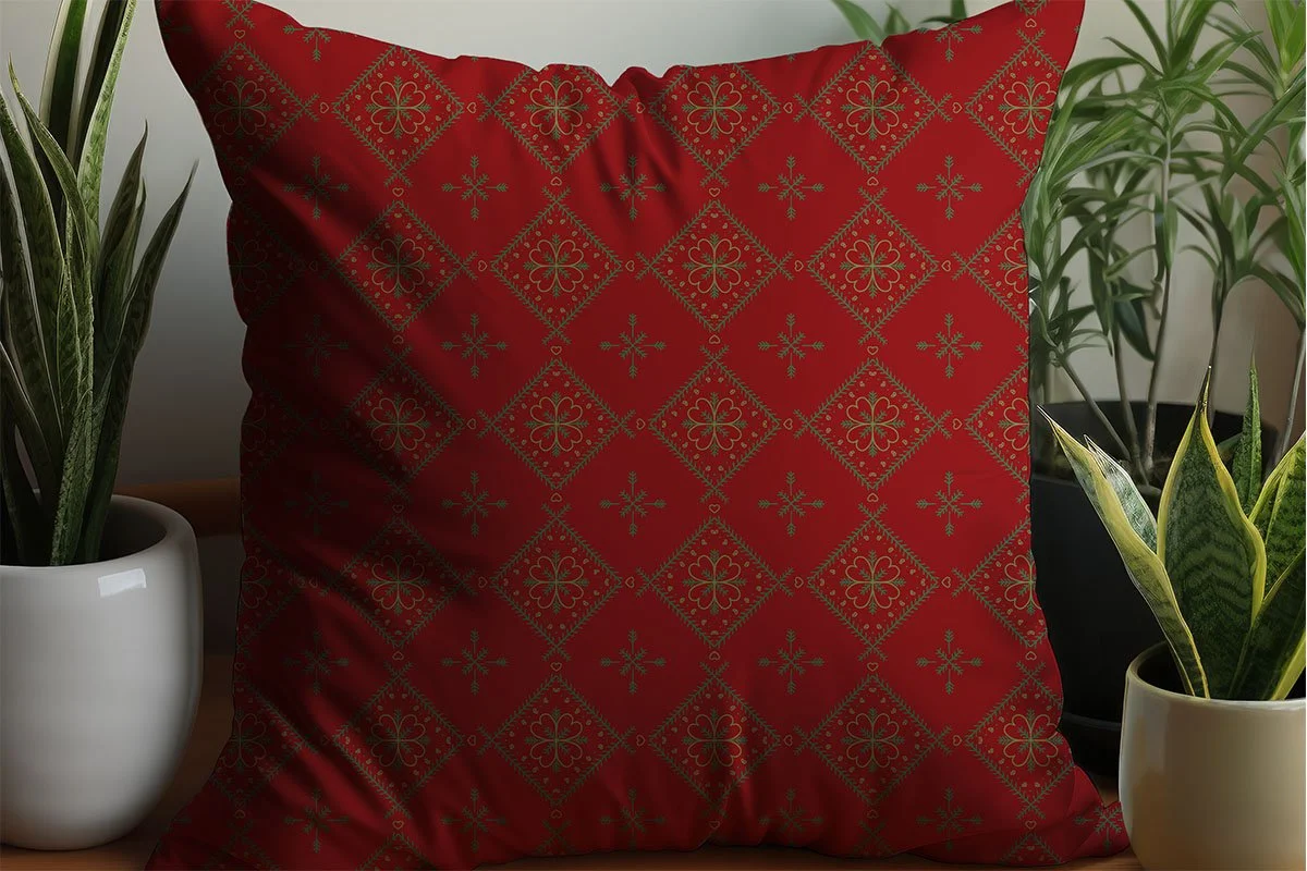 Red throw pillow with green Nordic-inspired hearts, evergreen branches and snowflake motifs, styled among houseplants.