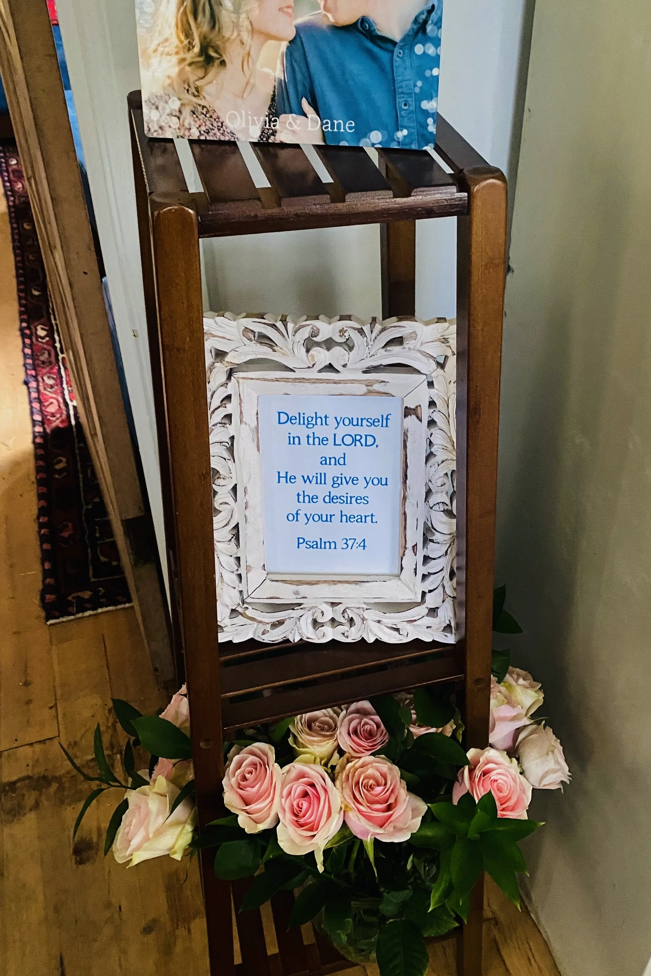 Wedding display featuring an engagement photograph, Psalm 37:4 framed artwork, and fresh roses arranged on a wooden stand.