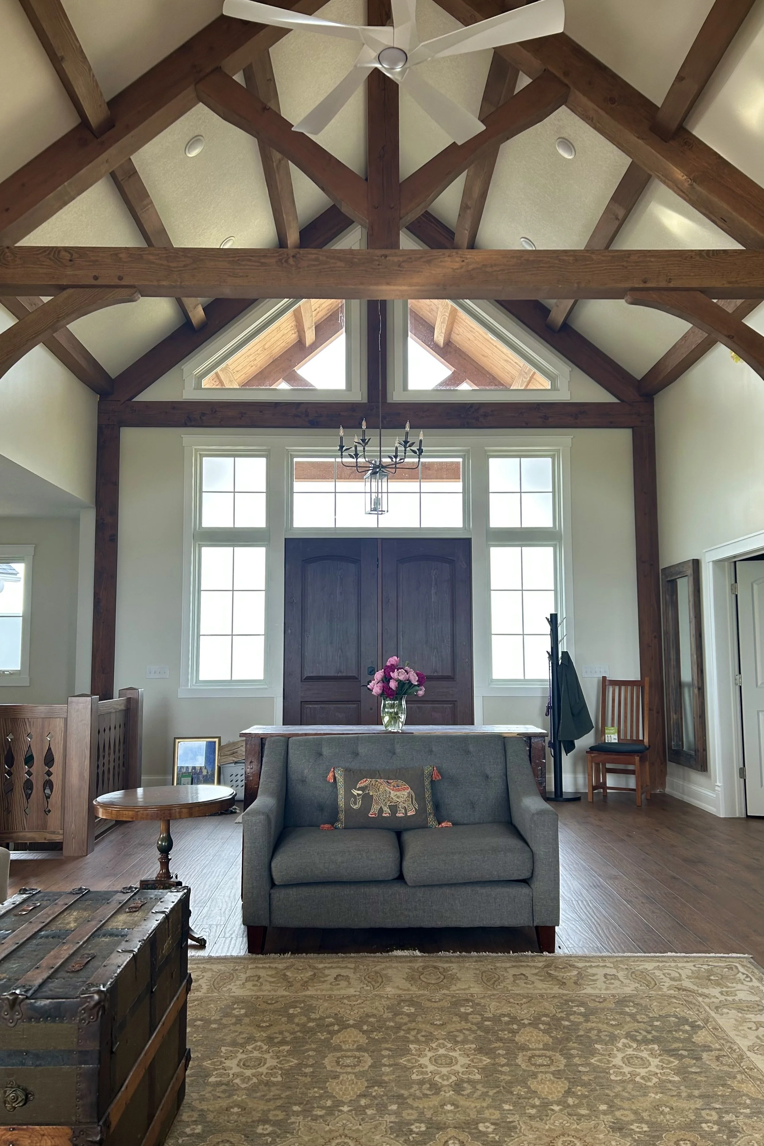 Wide view of a Minnesota farmhouse Great Room with exposed timber framing, tall windows, a gray sofa, antique trunk, patterned rug, flowers, and dark wood front doors.