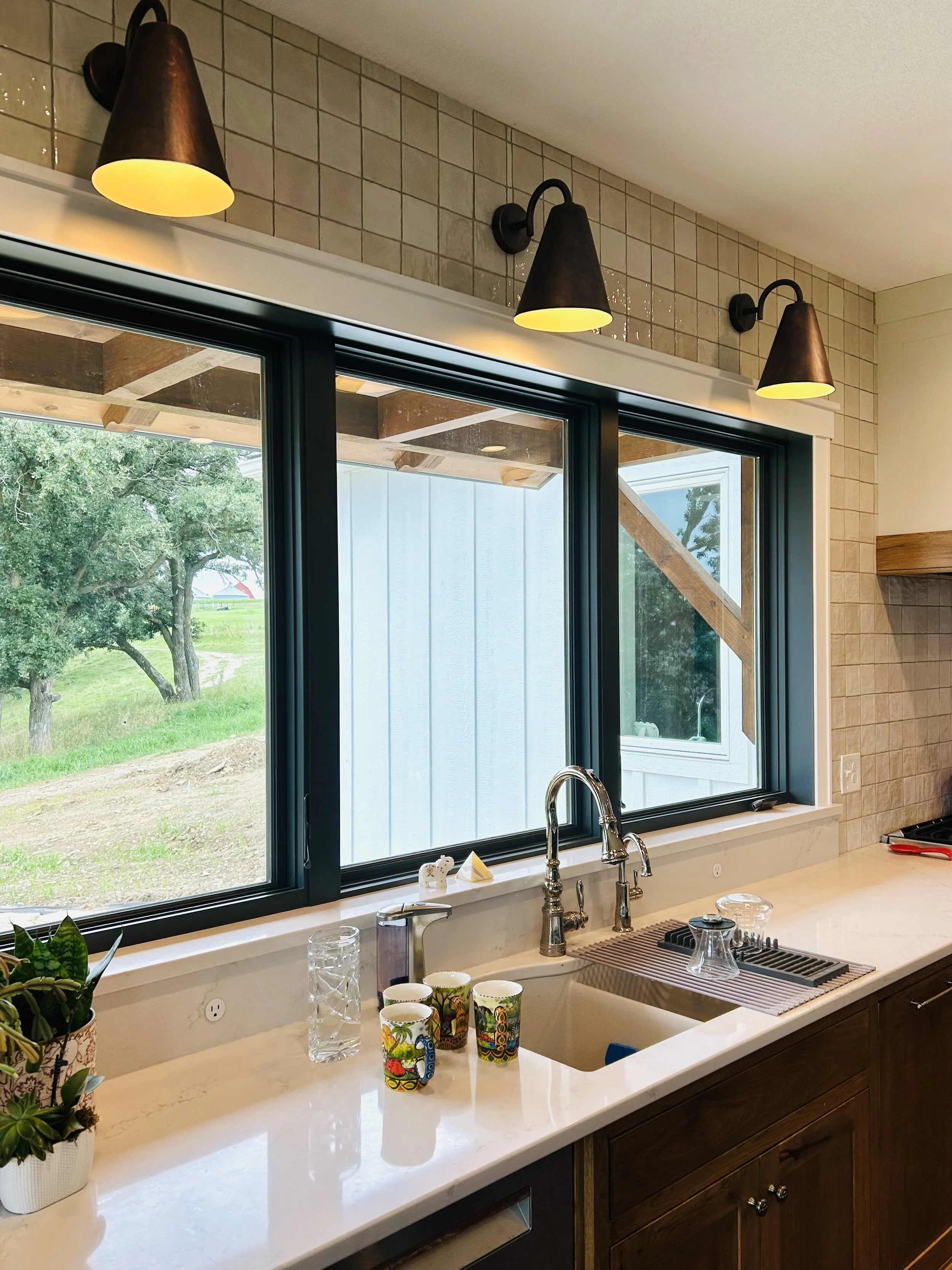 Handcrafted Moroccan sconces above a farmhouse kitchen sink overlooking rolling countryside, with travel mugs and meaningful keepsakes on the windowsill.