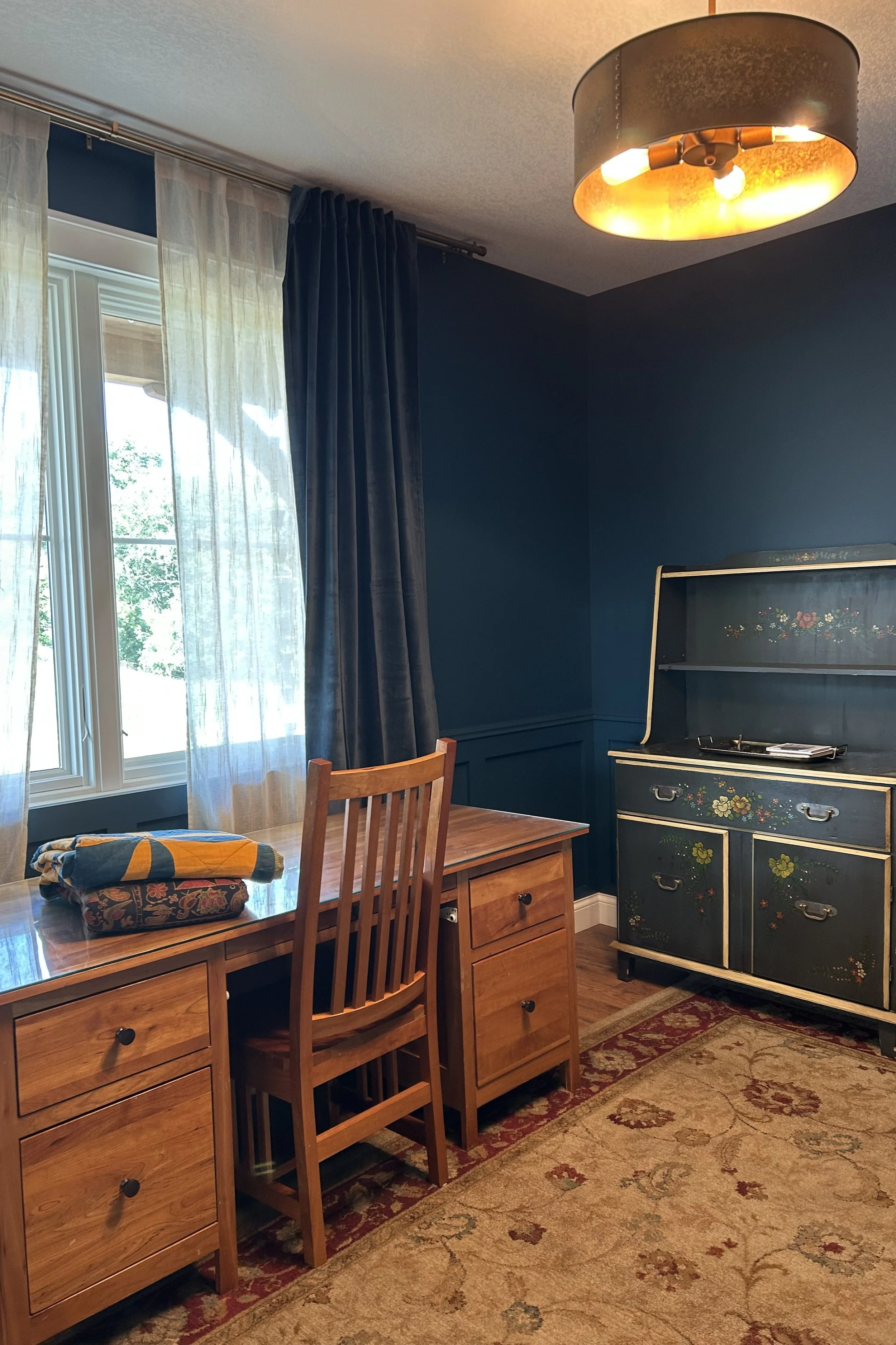 Traditional study with family heirloom quilt, folk-painted hutch, antique desk, upholstered chair, and patterned rug overlooking the Minnesota countryside.