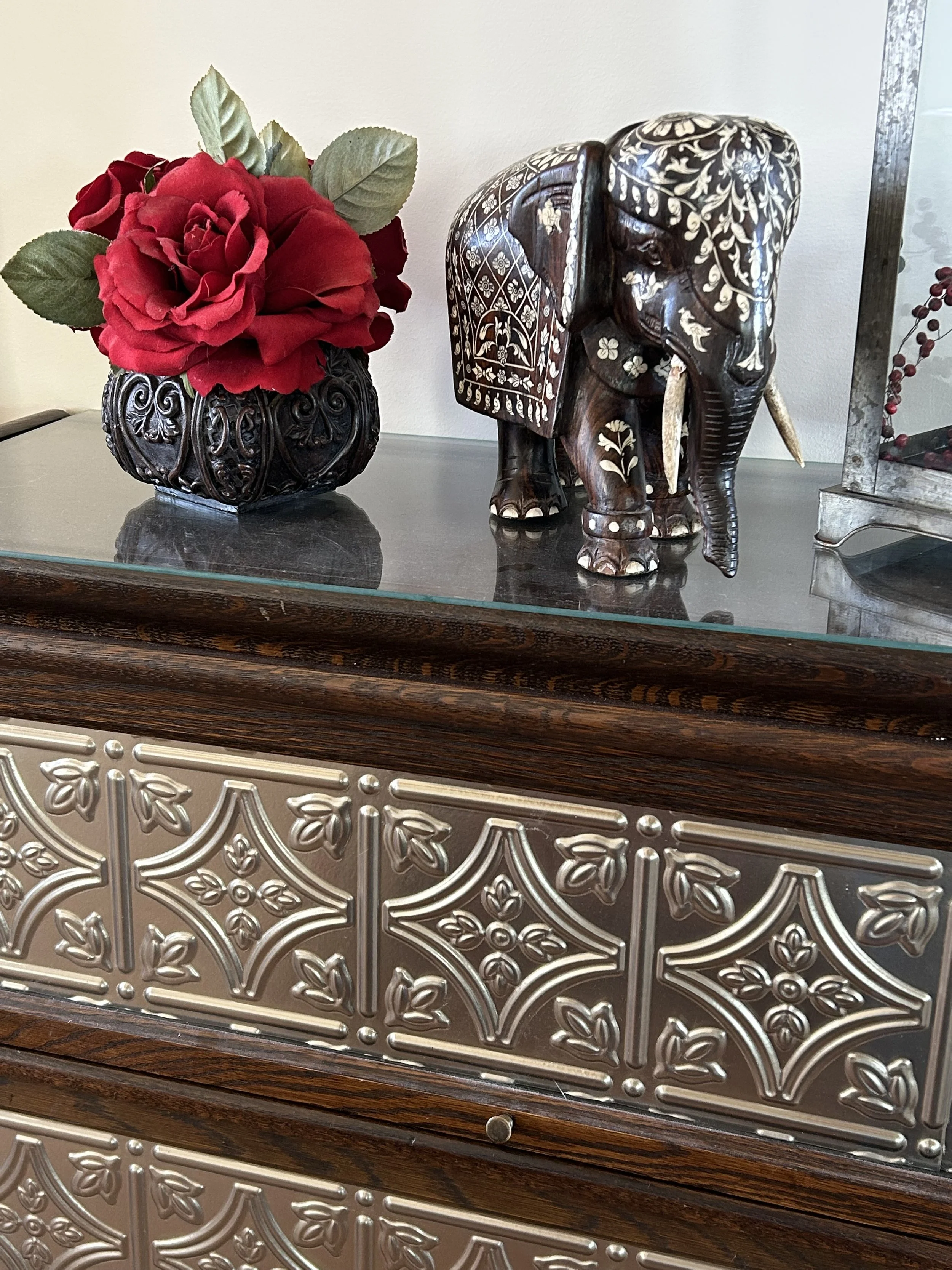 Carved dark wood elephant with inlaid floral details displayed beside deep red roses on an antique metal-front cabinet.