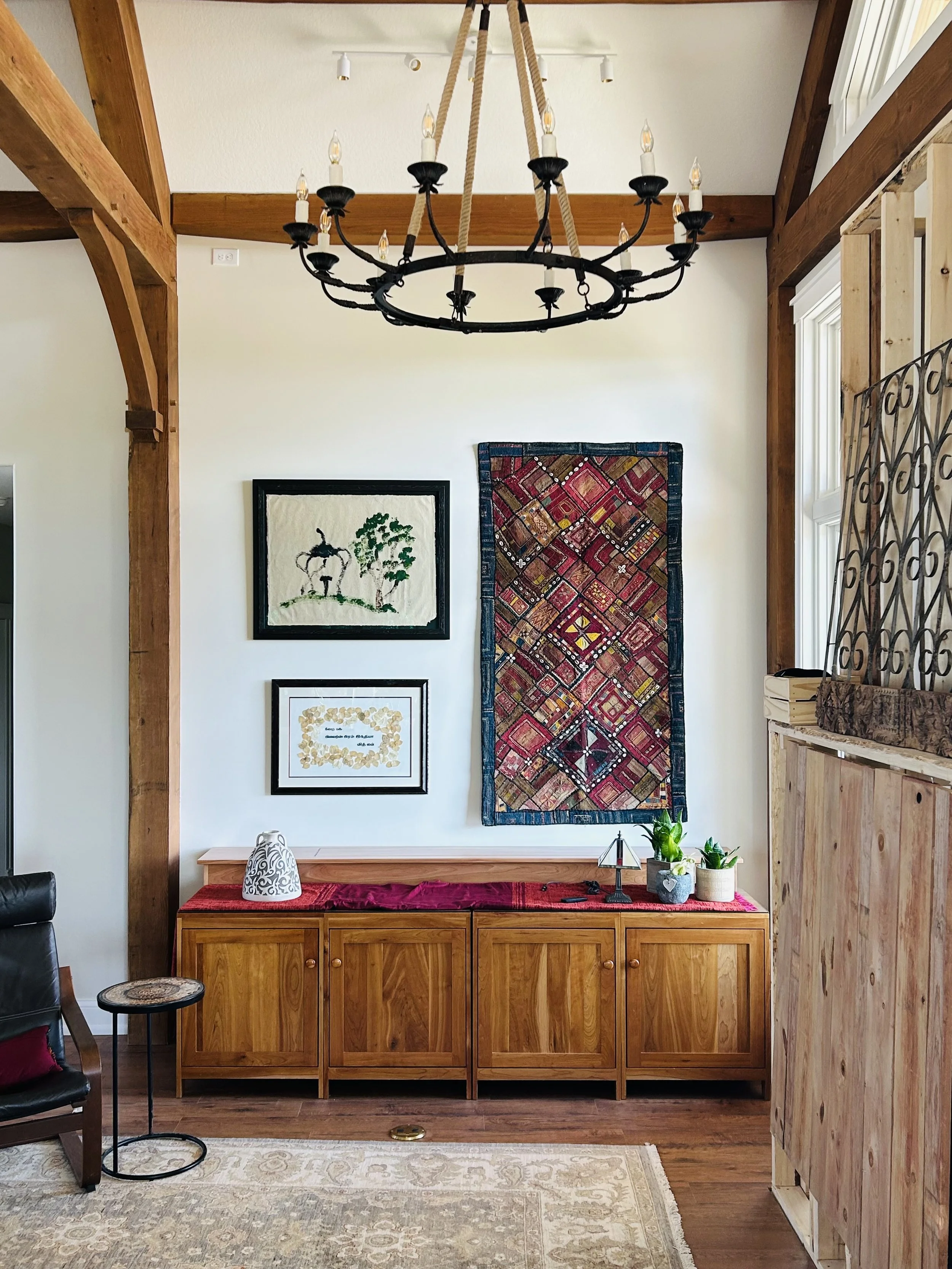 Timber-framed Great Room with handcrafted wood cabinets, framed artwork, an embroidered textile collected in India, plants, and meaningful objects displayed beneath a black iron chandelier.