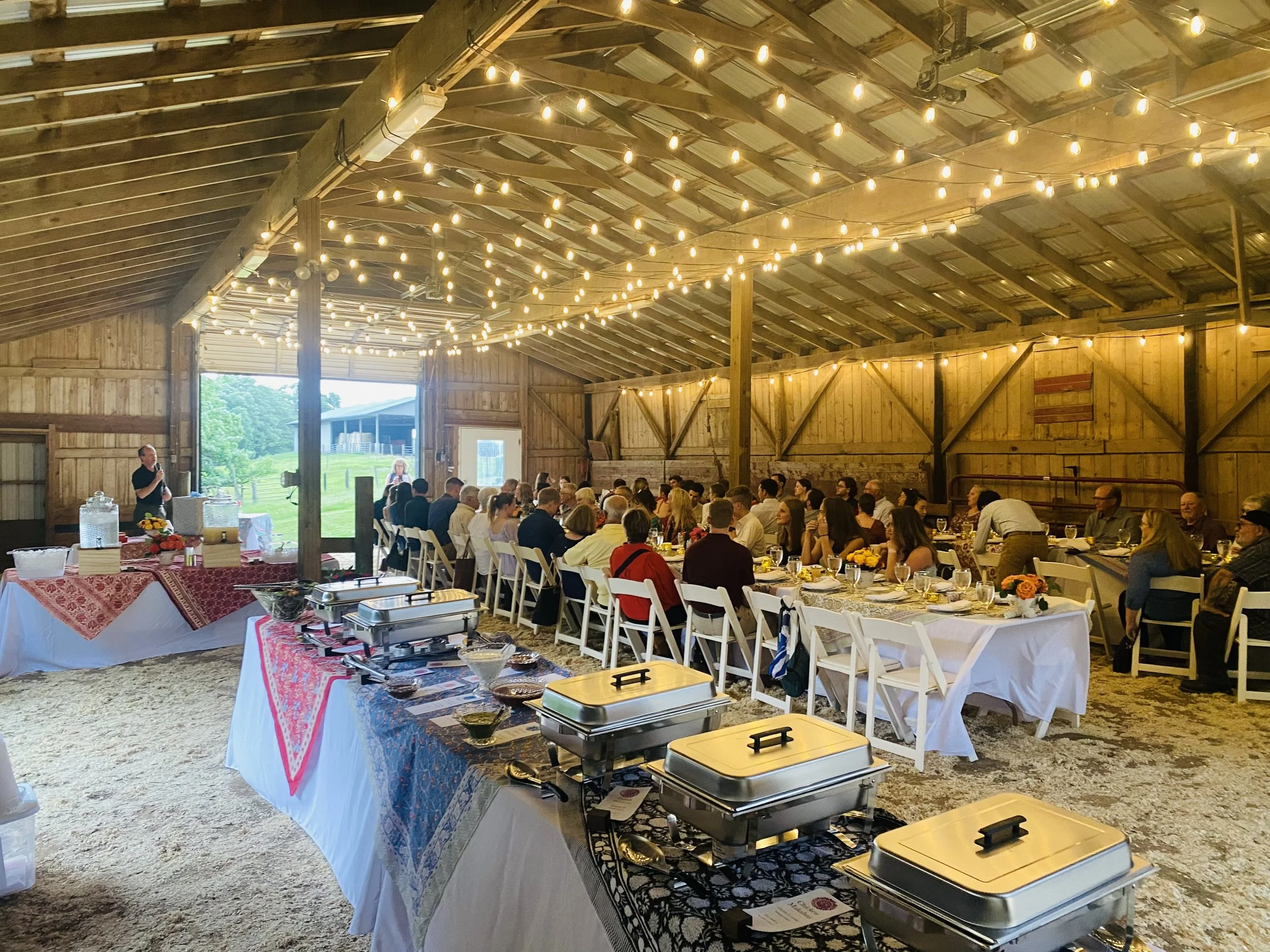 Guests gathered around long tables inside a rustic barn on our family farm for a groom's dinner beneath warm string lights.