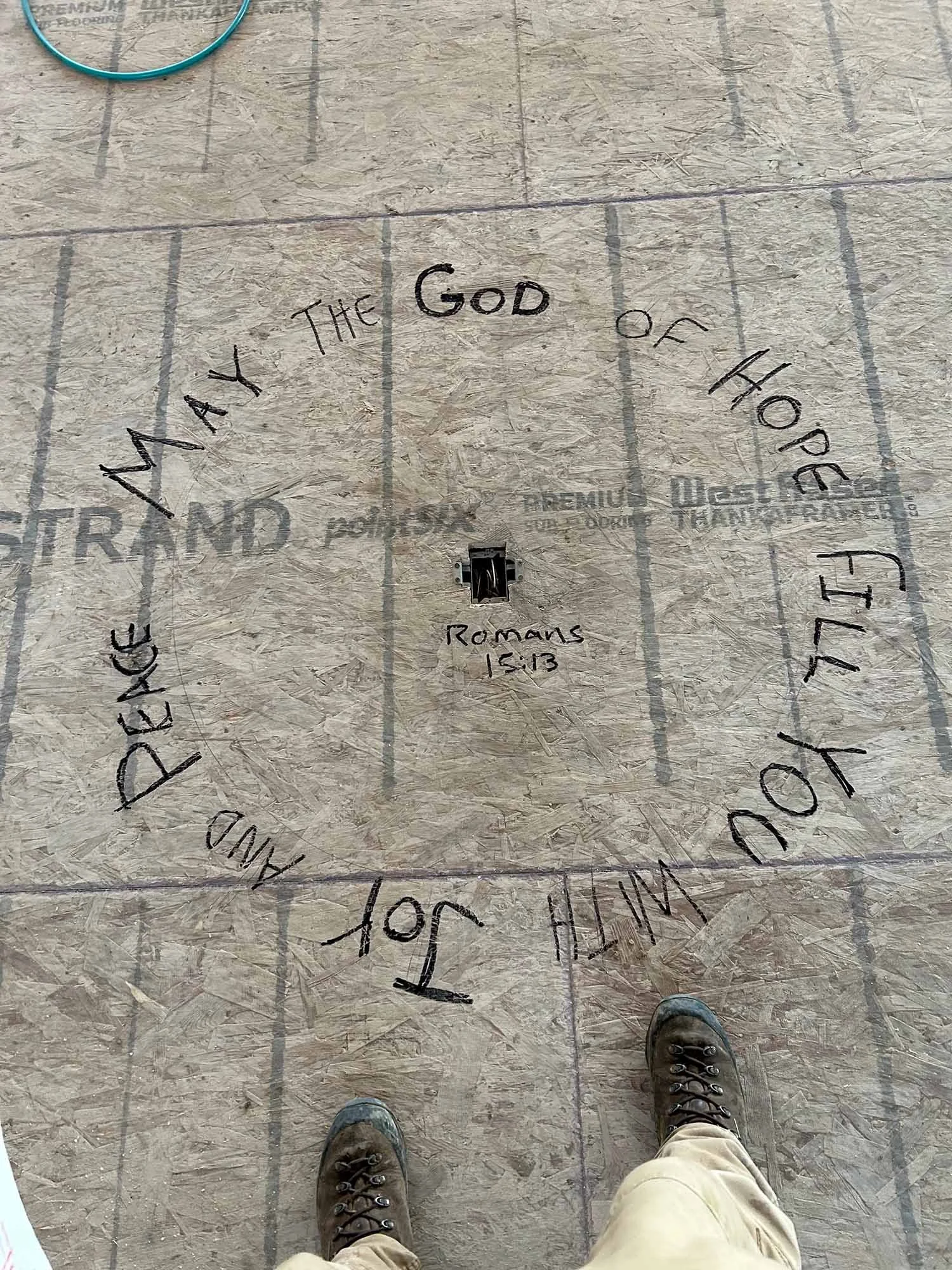 Romans 15:13 handwritten in a circle on the subfloor during construction, with the words about hope, joy and peace surrounding an electrical opening.
