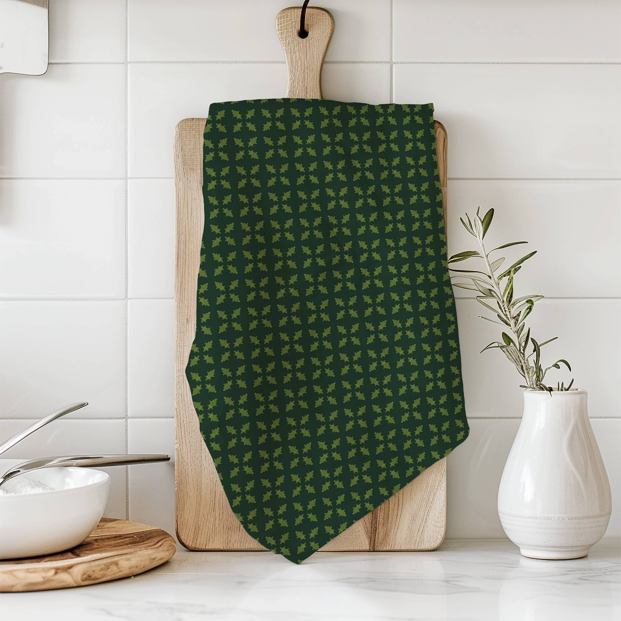 A green dish towel with lighter green leaf-shaped clusters hangs on a wooden cutting board in a kitchen. There is a white ceramic vase with a green plant on the right and a wooden cutting board with a white bowl and a silver spoon on the left.