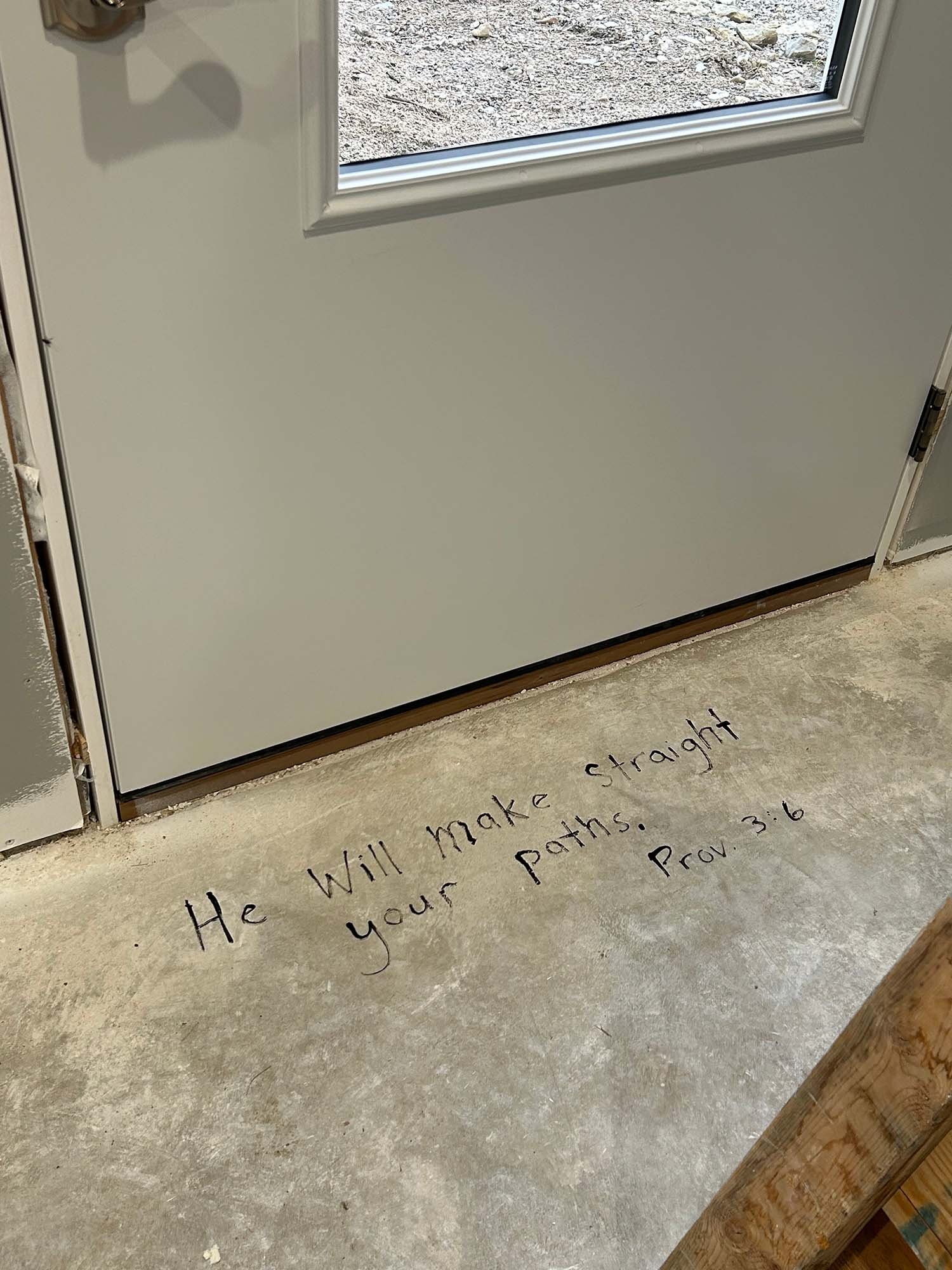 Proverbs 3:6, “He will make straight your paths,” handwritten on the unfinished floor beside a doorway during home construction.