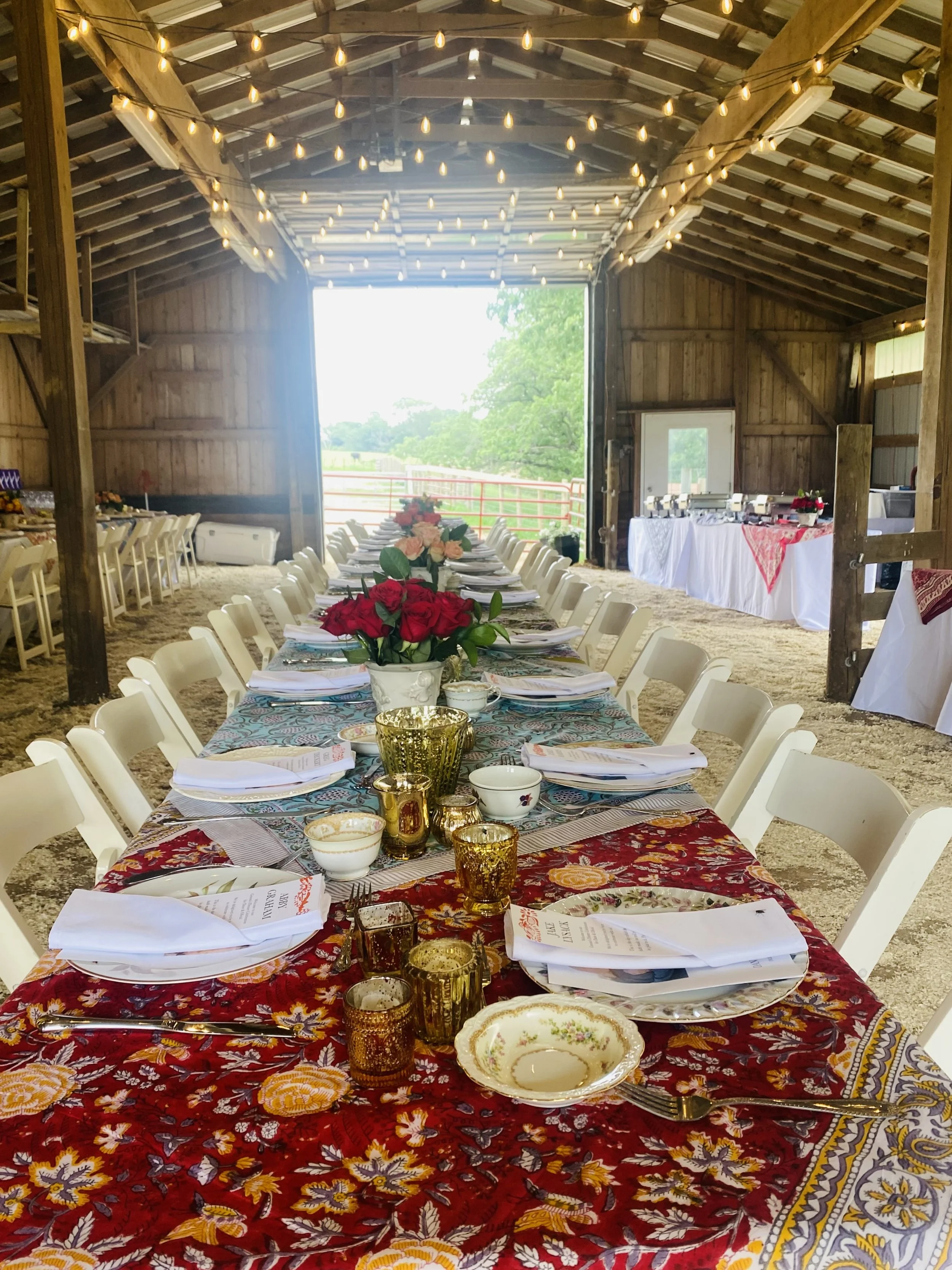 Long tables set inside a rustic barn with heirloom china, Indian textile table runners, fresh flower arrangements, candlelight, and white chairs prepared for a groom's dinner.