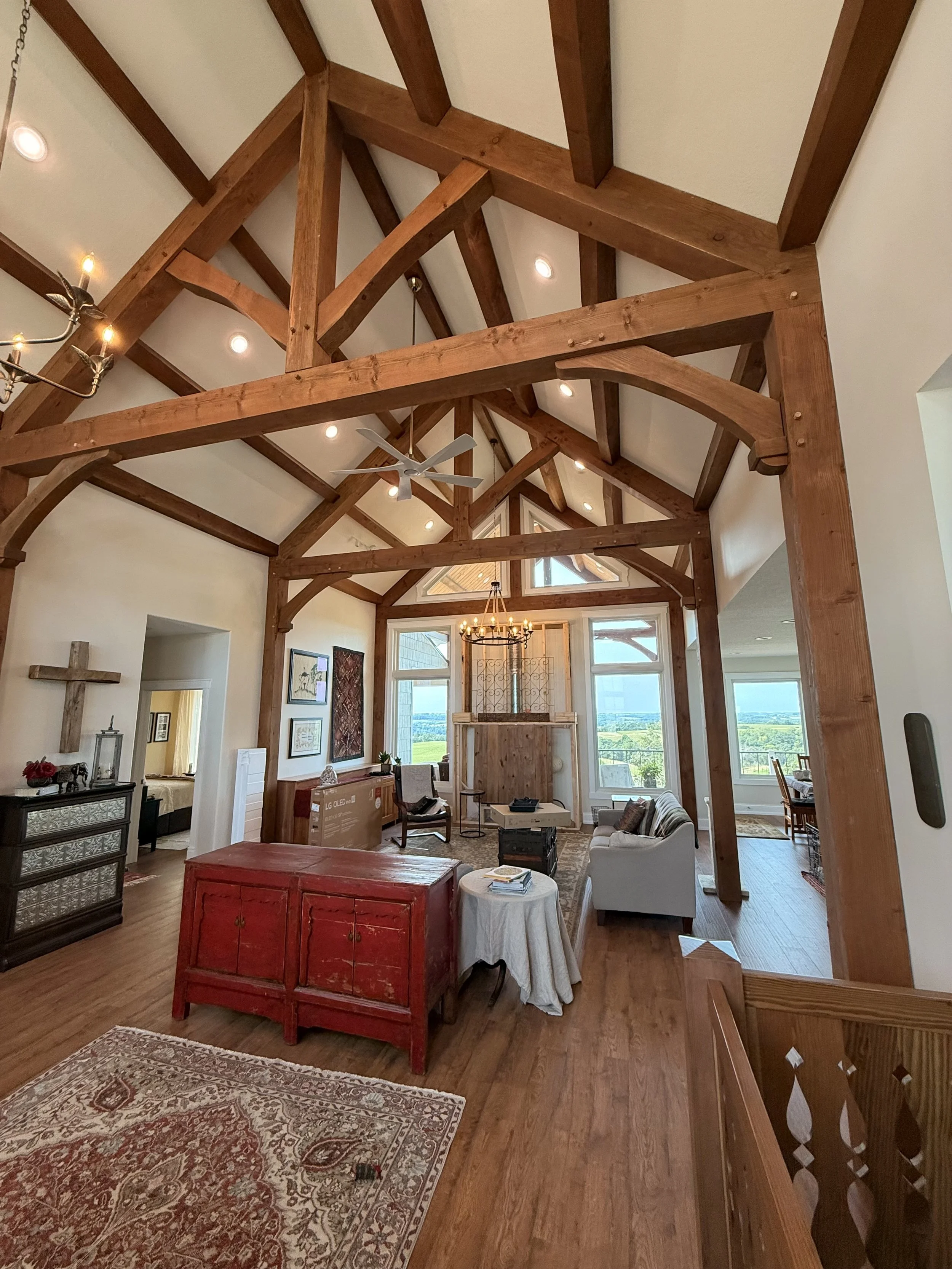 Exposed timber-frame beams and vaulted ceiling in a great room with collected furnishings, patterned rugs and views across the surrounding countryside.