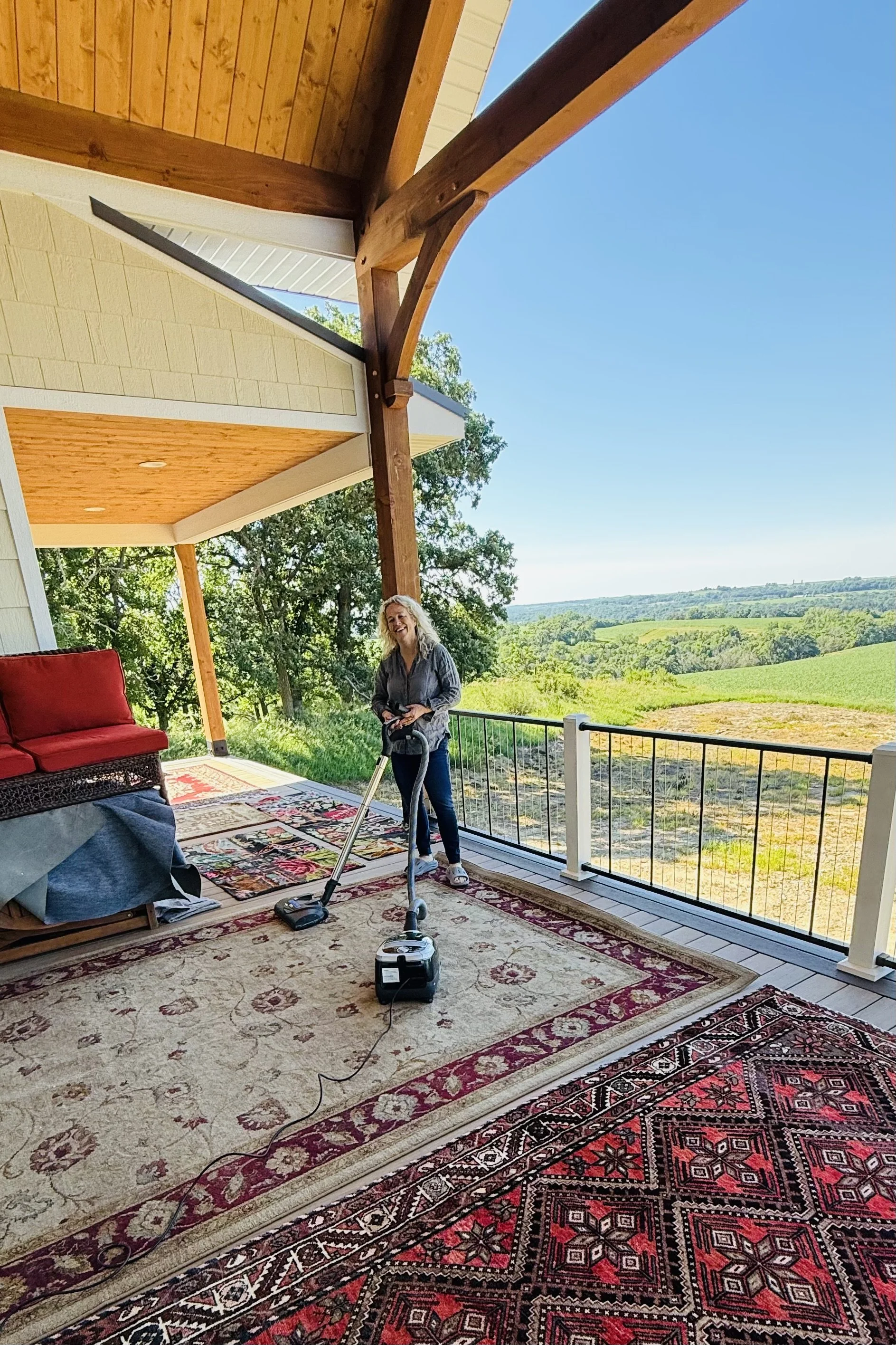Cleaning and airing rugs on a covered back deck overlooking rolling farmland and wooded hills.