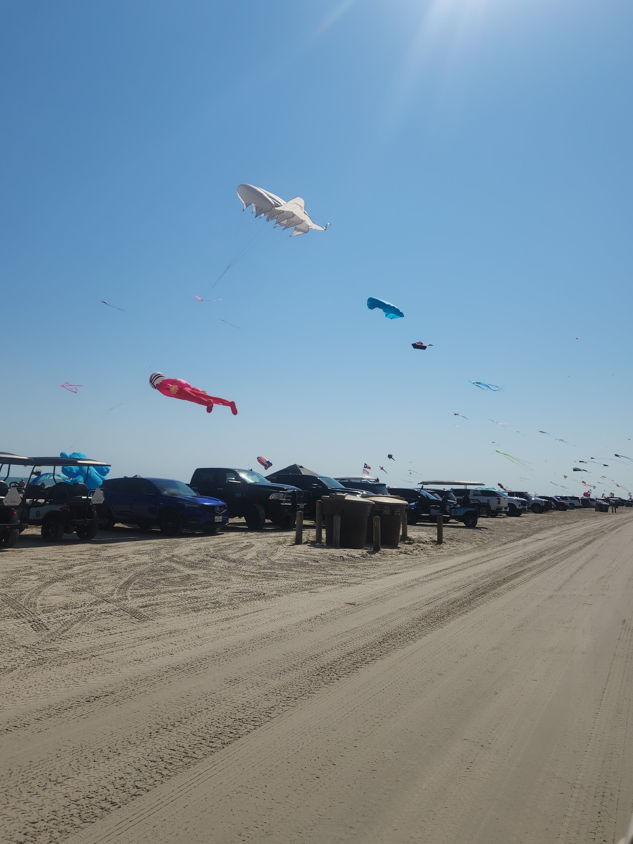 Beach scene with colorful kites flying above parked cars and buggies on the sand.