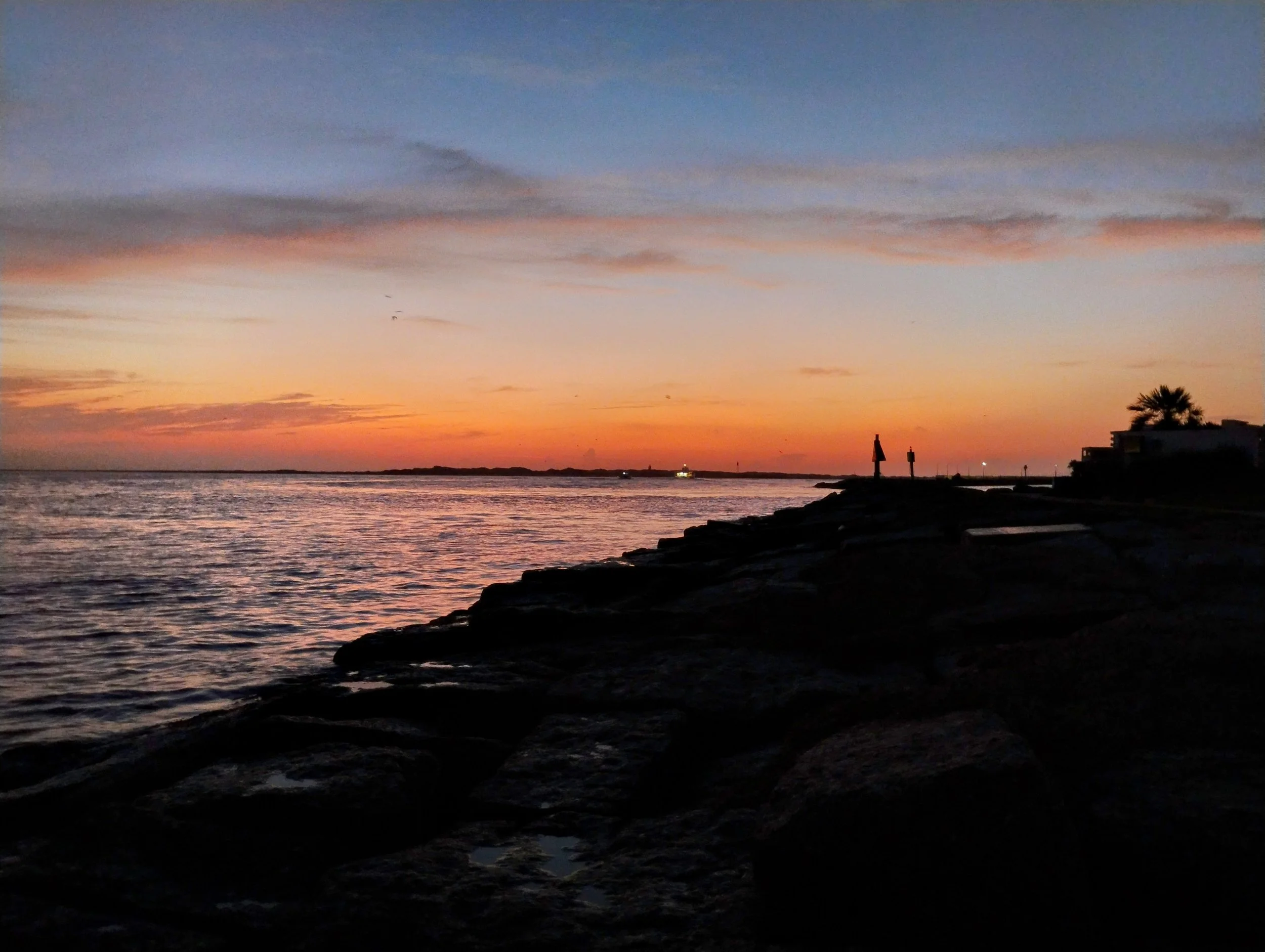 Sunset over a body of water with orange and pink hues in the sky, silhouetted rocks in the foreground, and small structures and palm trees on the shoreline. Port Aransas, TX