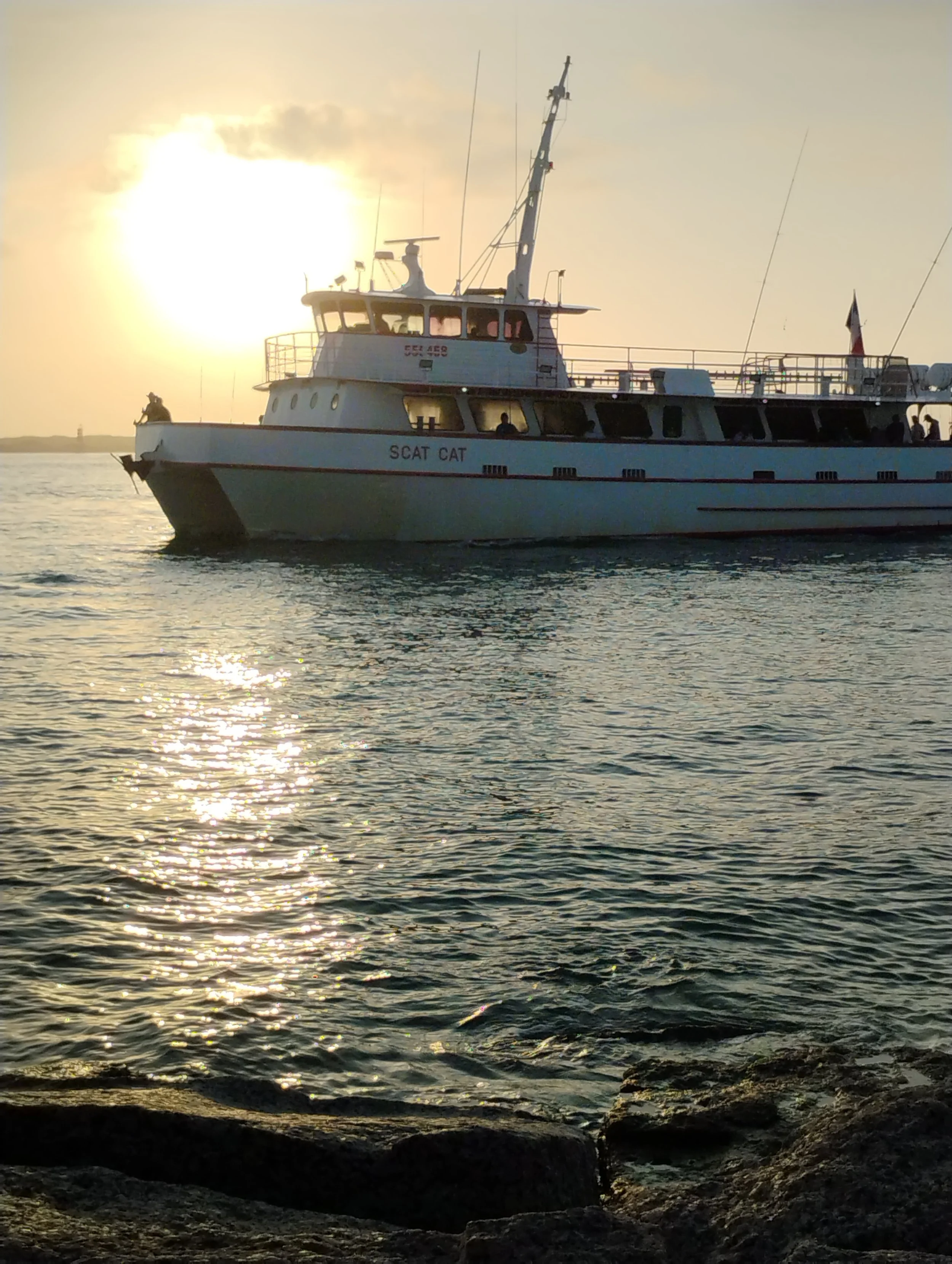 A white boat named 'SCAT CAT' sailing on water during sunset, with the sun low on the horizon and reflections on the water surface. Port Aransas, TX