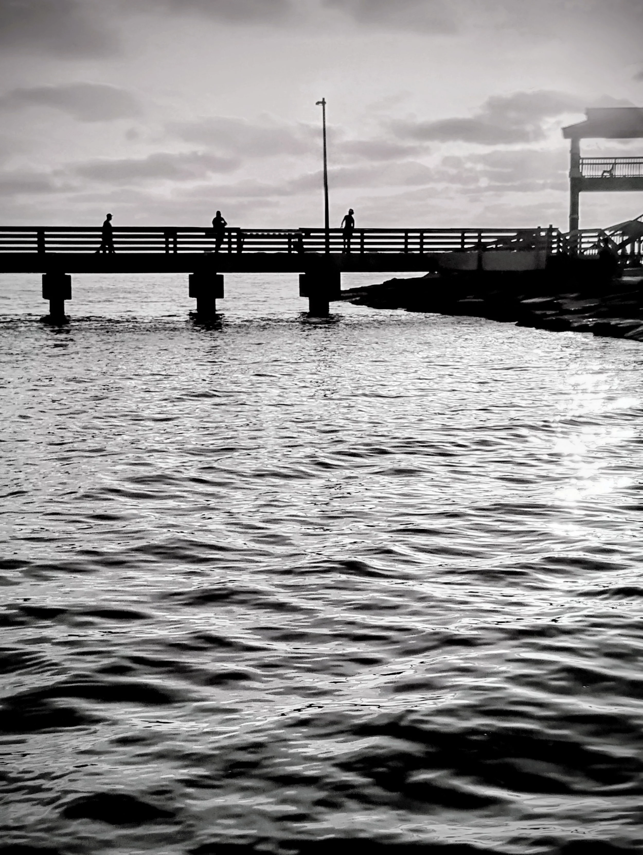 Silhouette of people walking on a pier over water, with a cloudy sky in the background, in black and white. Port Aransas, TX