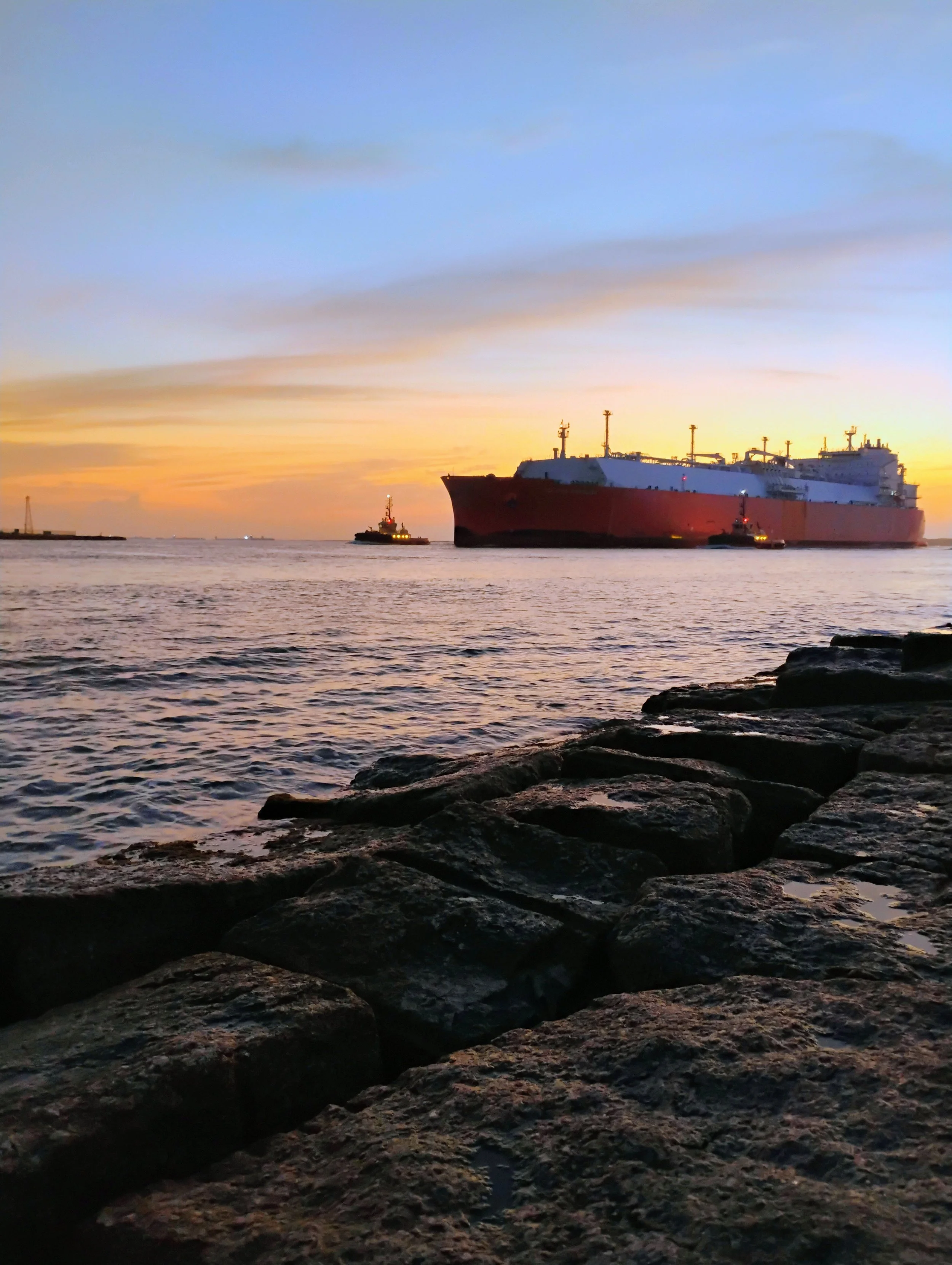 Large cargo ship being guided by tugboats during sunset near rocky shoreline. Port Aransas, TX