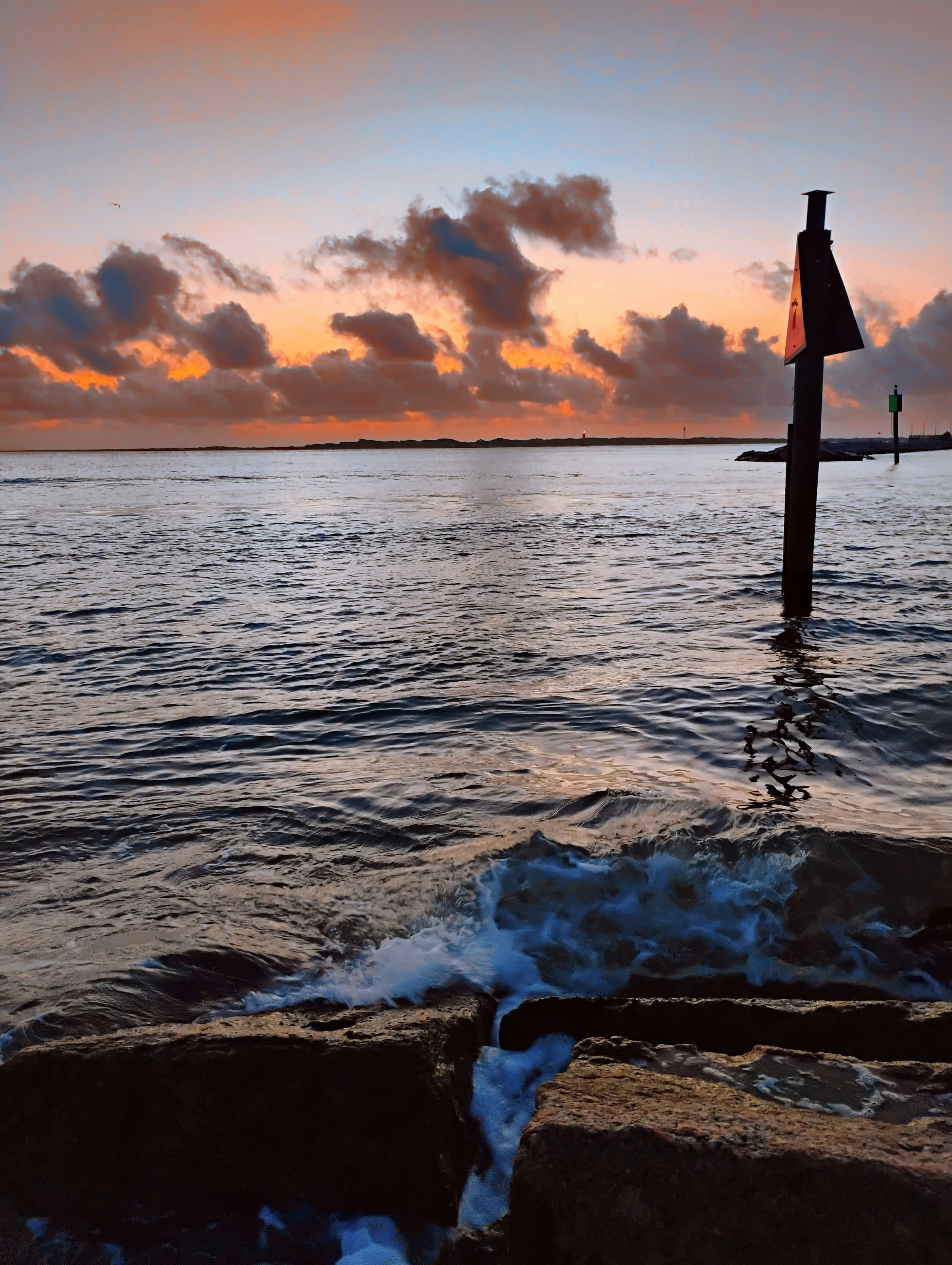 Sunset over a calm ocean with a partly cloudy sky, rocks in the foreground with waves gently crashing, a signpost in the water, and a distant shoreline. Port Aransas, TX