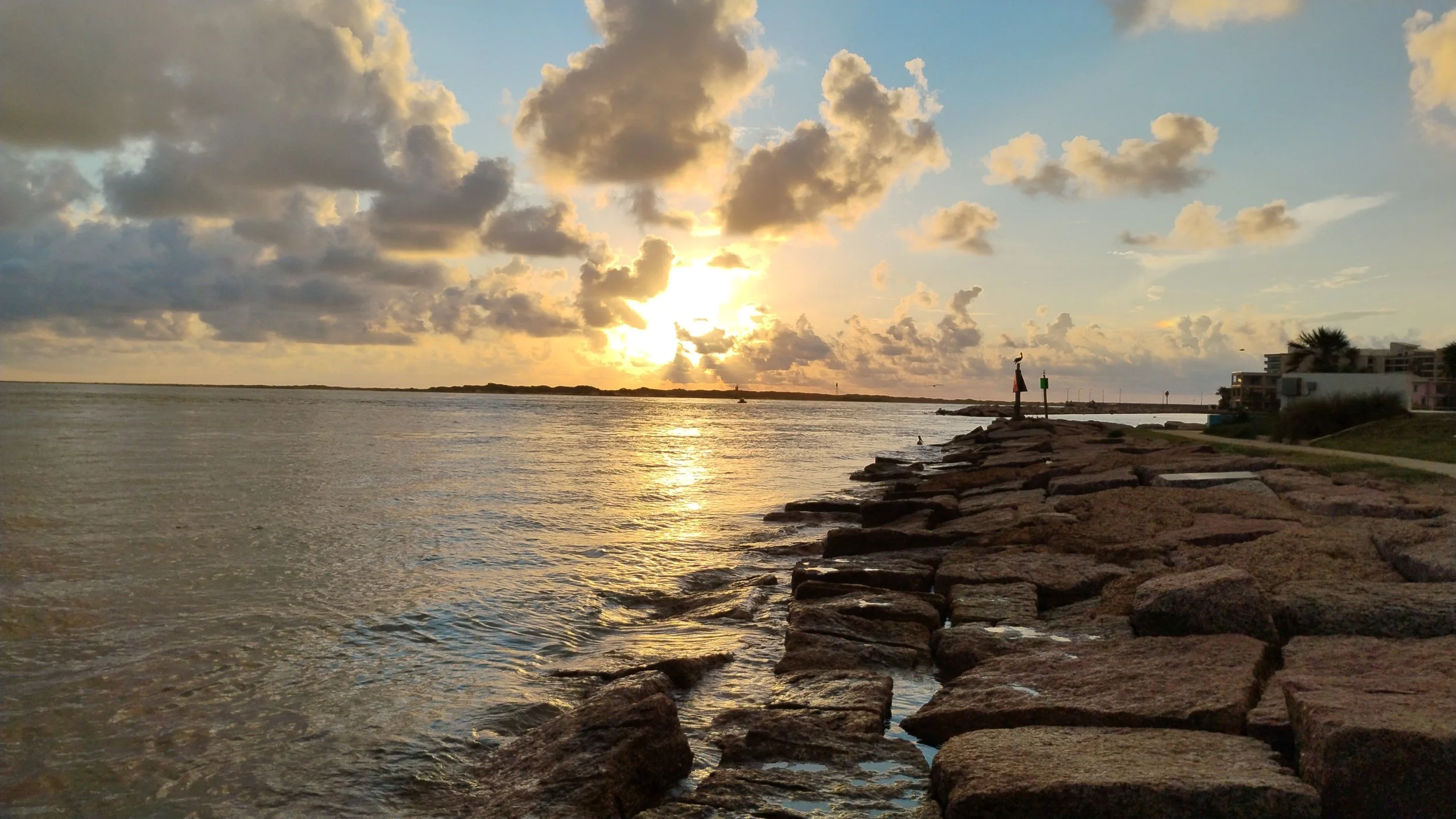 Sunset over a body of water with a rocky shoreline and clouds in the sky. Port Aransas, TX