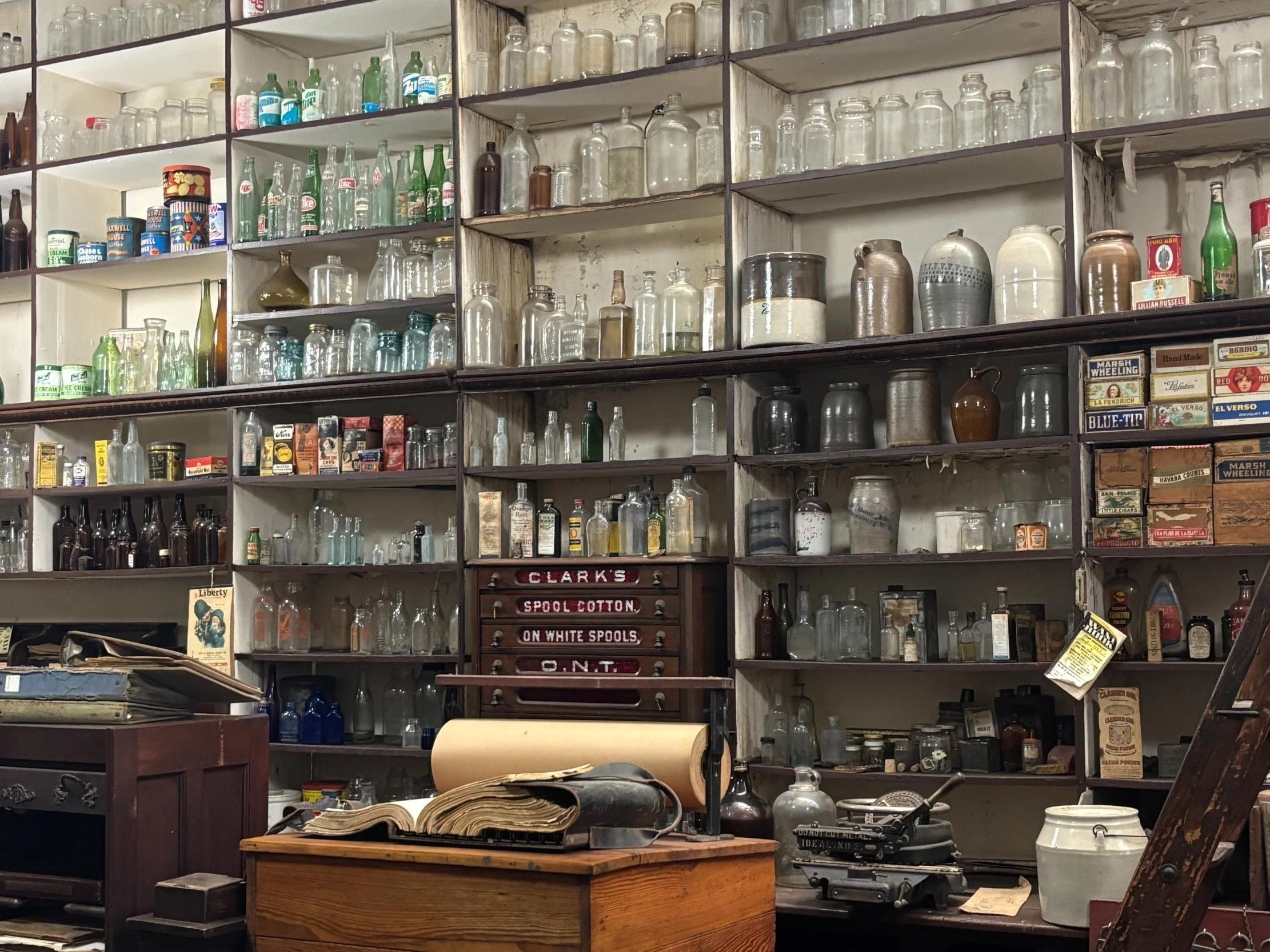 A collection of vintage glass bottles and jars on shelves in an antique shop, with old advertisements and typewriters nearby.