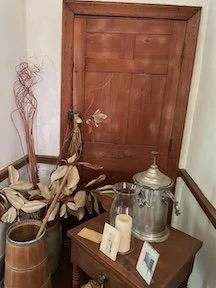 Decorative corner with a wooden door, dried leaves in a vase, a glass lantern, a candle, and a framed photo on a small table.