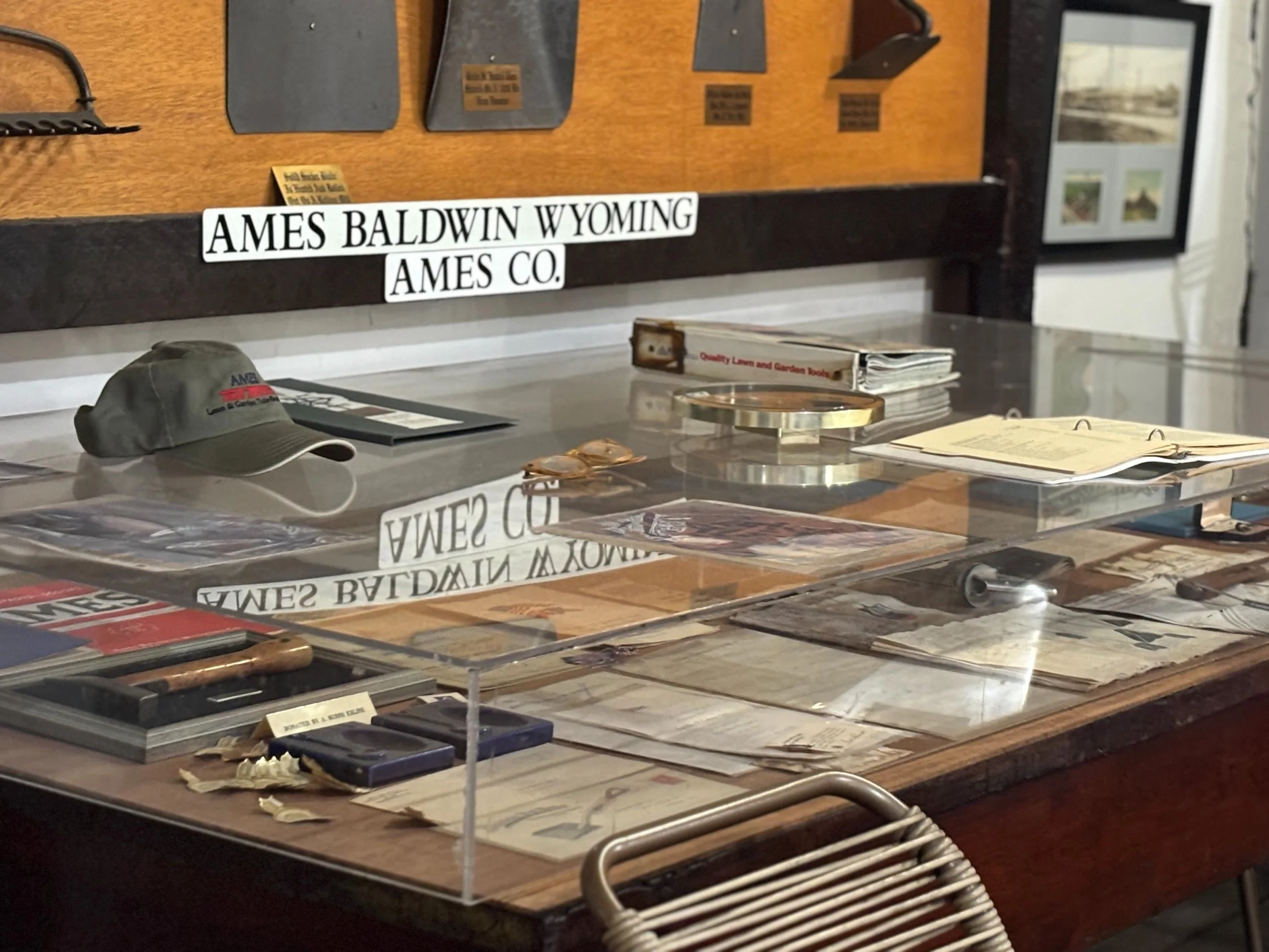 Historical display case with papers, a cap, a book, and various memorabilia, with a sign reading "Ames Baldwin Wyoming Ames Co." in the background.