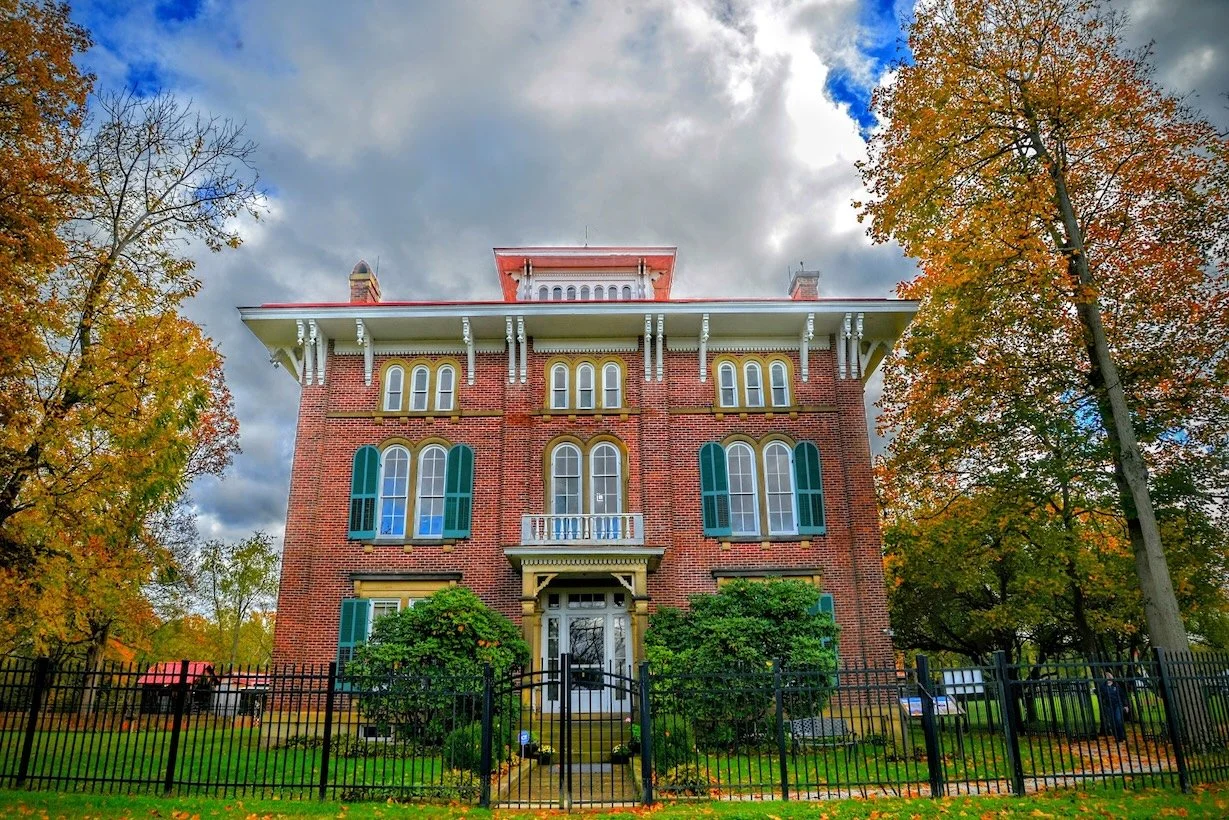 A large, historic brick mansion with green shutters, a white balcony, and a fenced front yard surrounded by autumn trees.