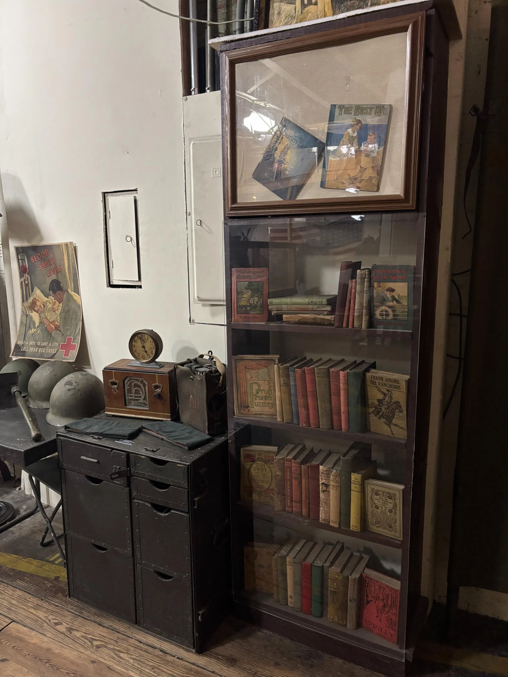 A glass-fronted wooden bookcase filled with vintage books, a small clock, and various decorative items sit on a hardwood floor next to a black metal drawer organizer. There are military helmets and a vintage radio on the left, and some military memorabilia on the wall.