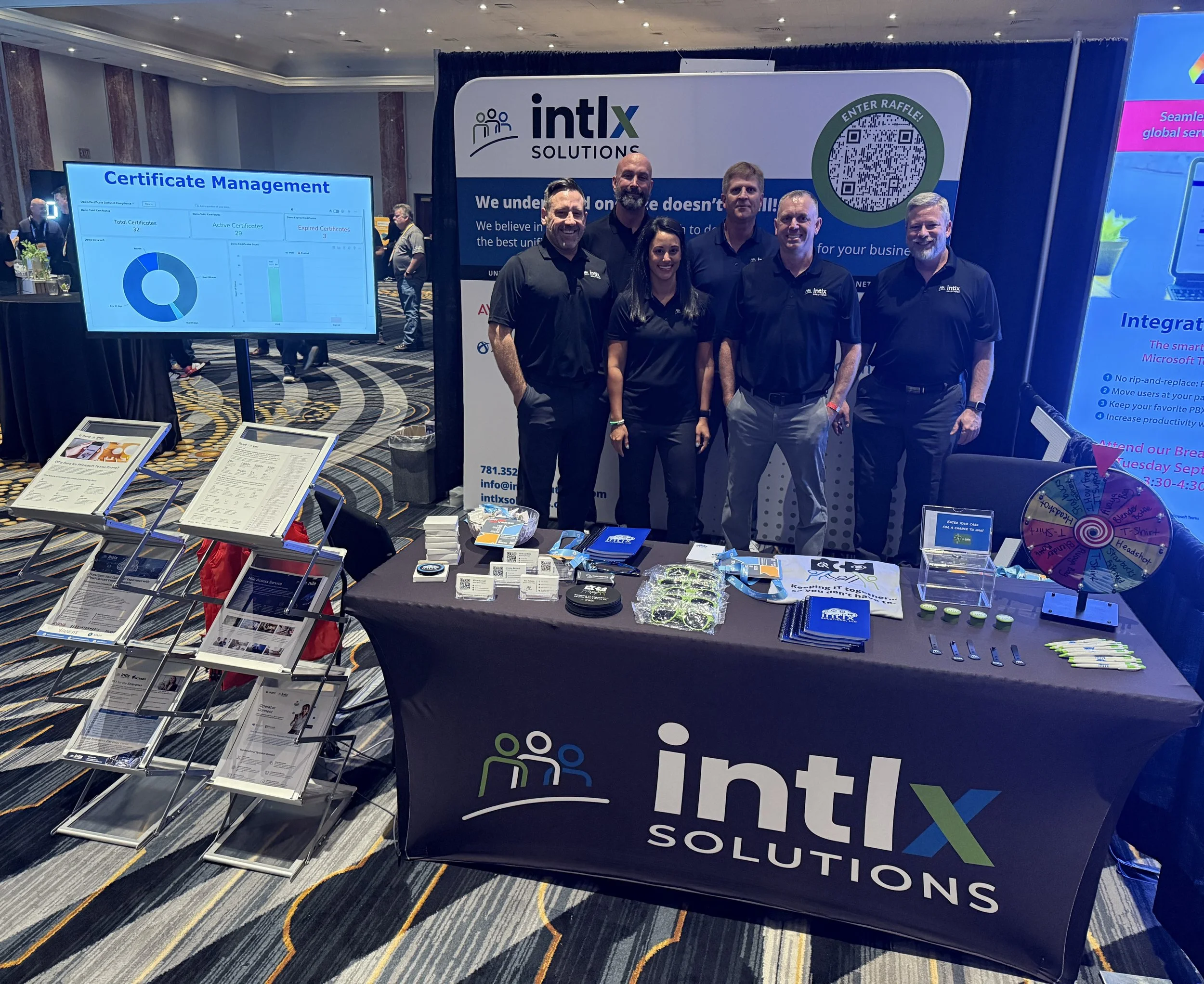 A group of five people standing behind a booth for Intlx Solutions at a conference or trade show. The booth displays a table with branded materials, brochures, and a spinning prize wheel. Behind them, a large banner and a digital screen show company information and graphics related to certificate management. The booth appears professional and well-organized, with other booths and attendees visible in the background.