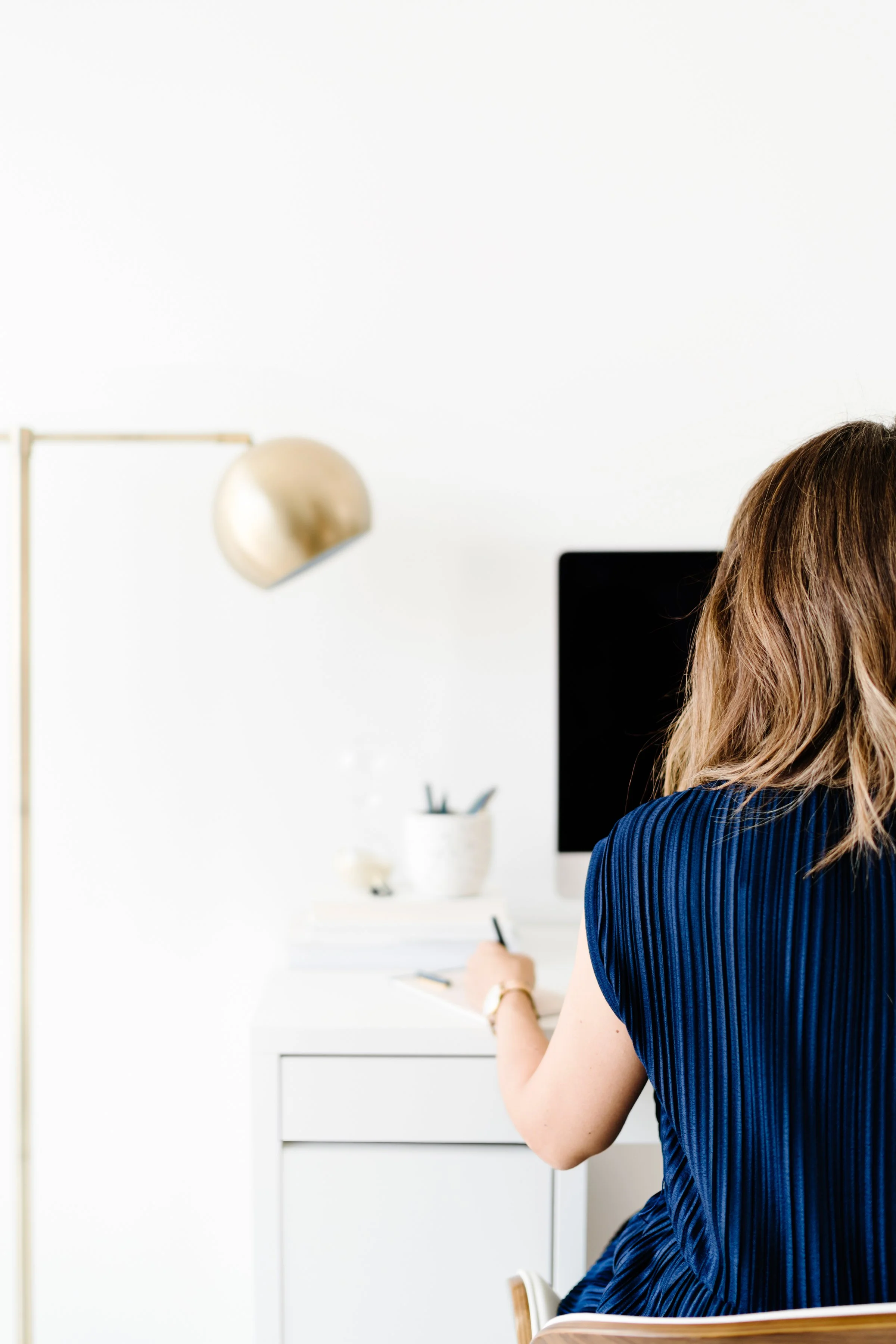 A woman working on a laptop at a desk in a bright office with plants, a notebook, a mug, and a lamp.