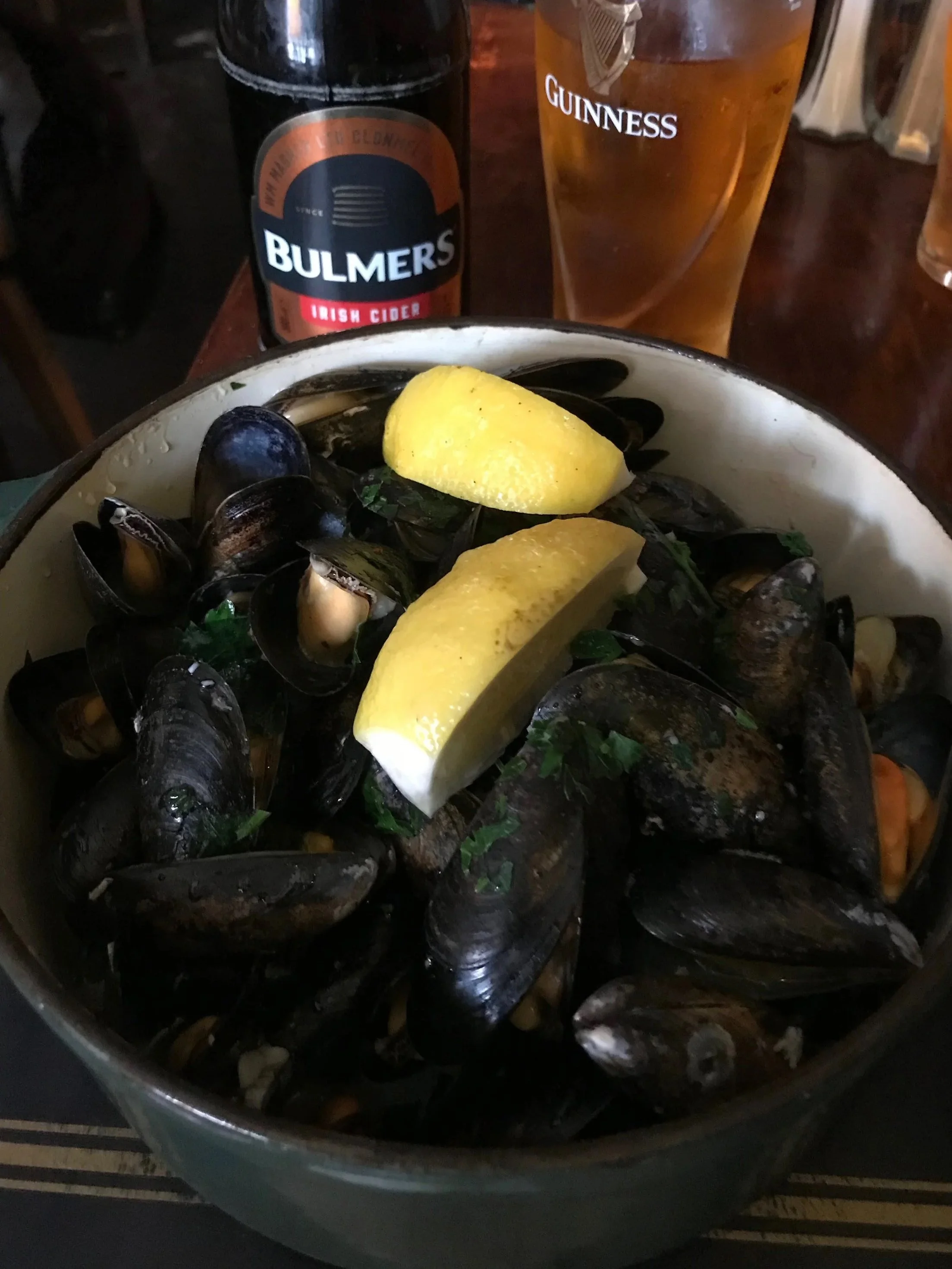 A bowl of cooked mussels garnished with lemon wedges and herbs, with a bottle of Irish cider and a pint of Guinness beer in the background.