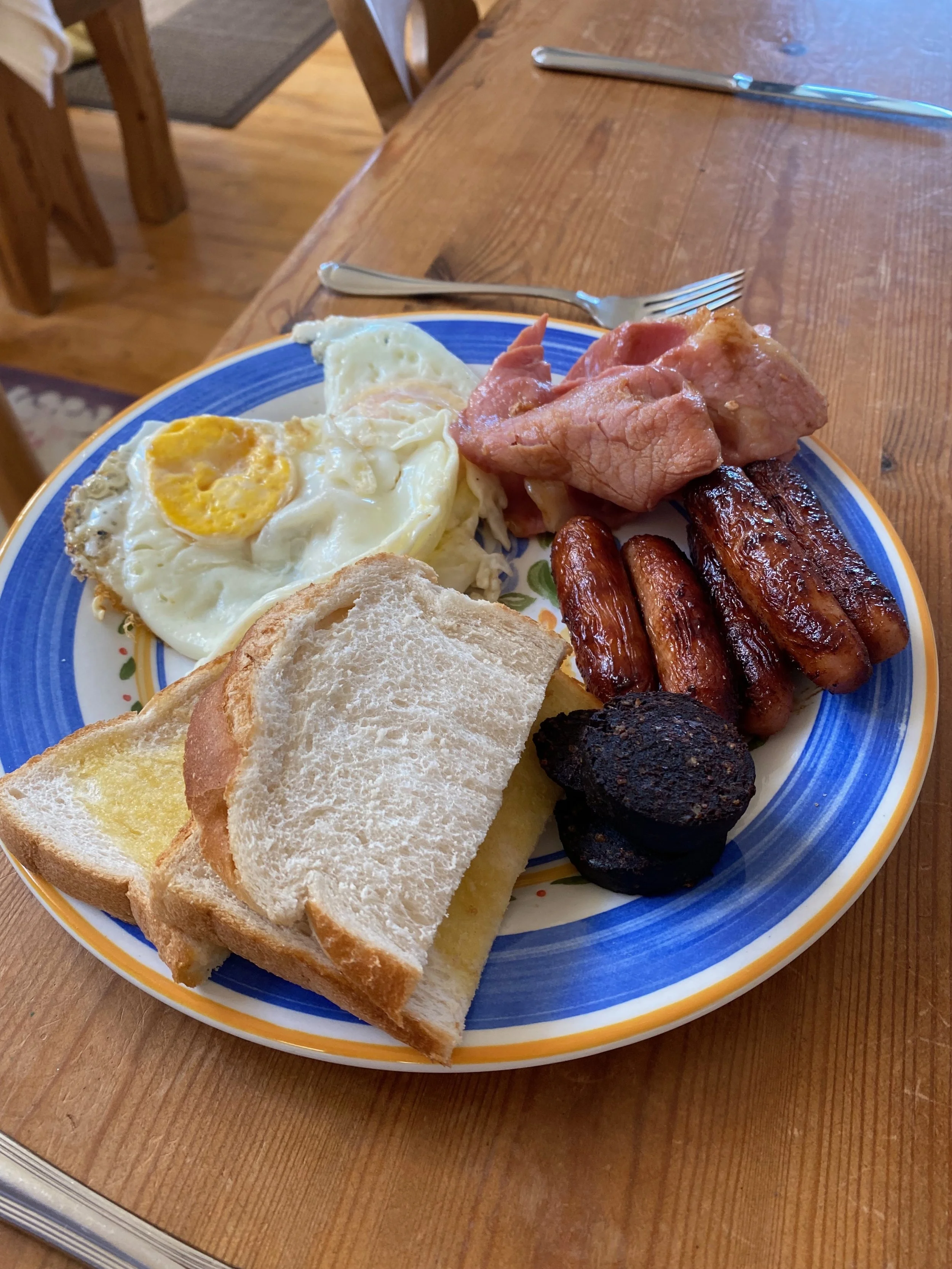 A breakfast plate with fried eggs, toast, bacon, sausage links, black pudding, and grilled ham on a blue-rimmed plate.