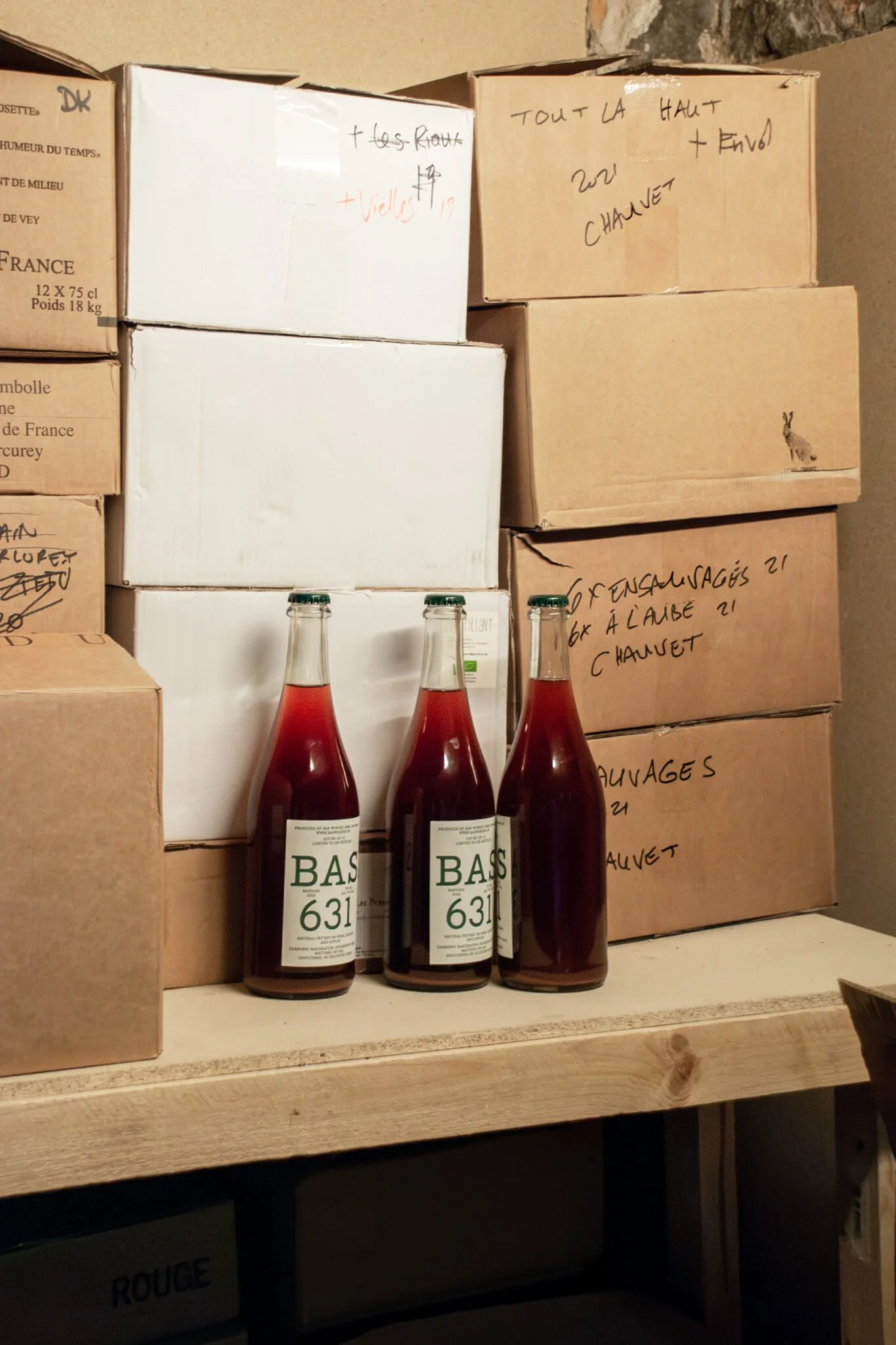 Multiple cardboard boxes stacked on a shelf with handwritten notes and labels, with three bottles of a beverage in front of the boxes.