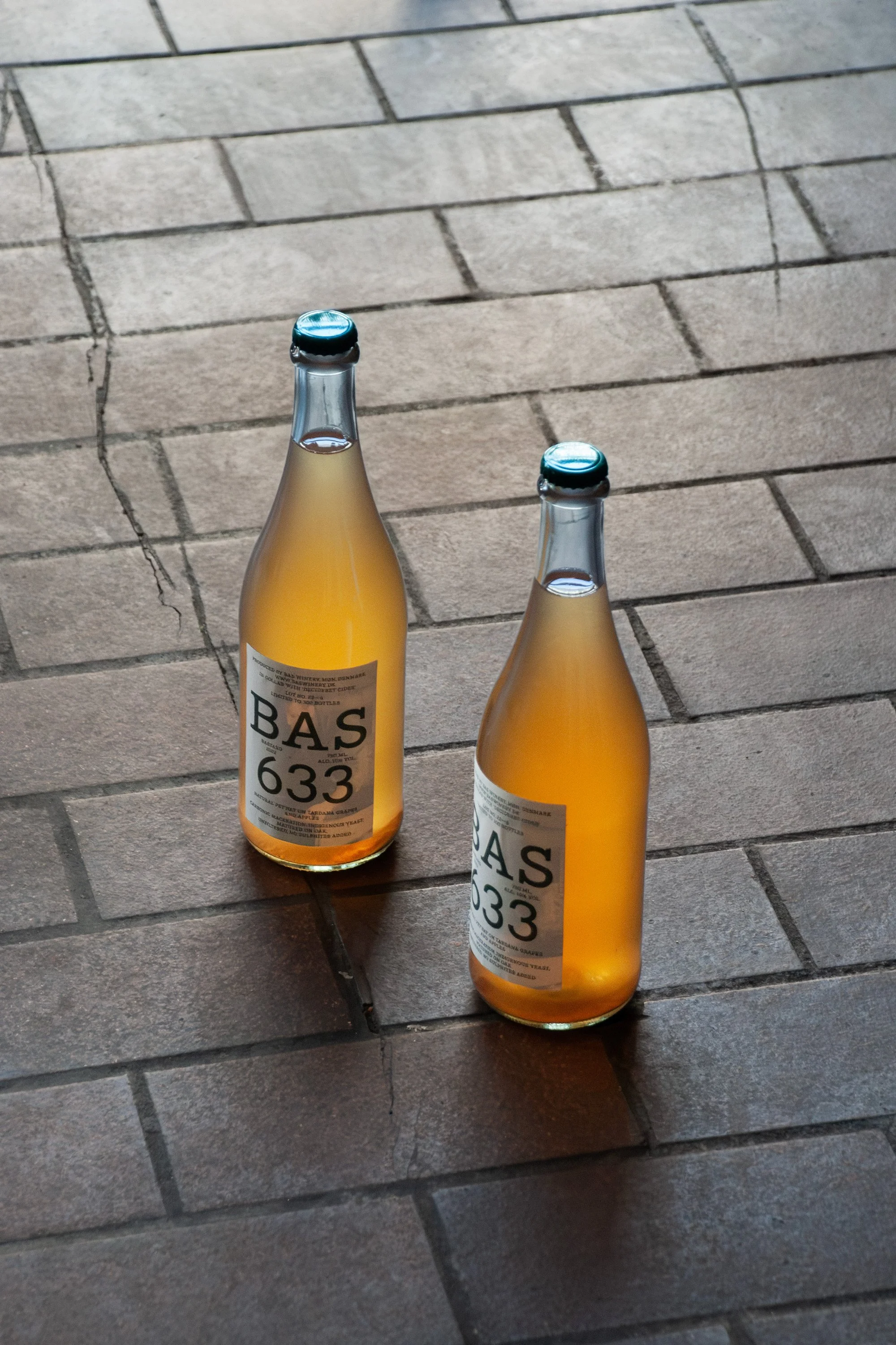 Two bottles of wine with light amber color on a tiled floor, one slightly behind the other, with labels that have large text and a golden hue.