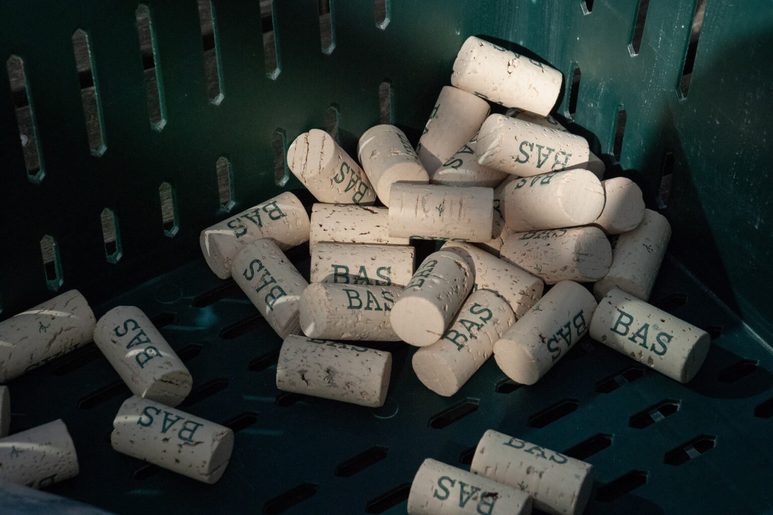 A green plastic basket containing used white wine corks with the word 'BAS' printed on them.
