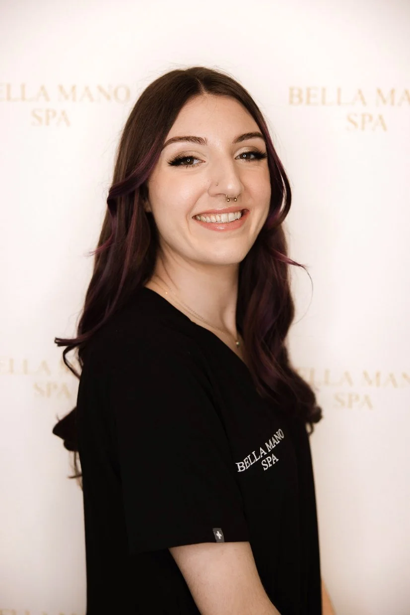 A smiling woman with long, wavy purple hair and septum piercing wearing a black uniform standing in a modern, bright reception area with a white marble counter and a sparkling chandelier overhead.