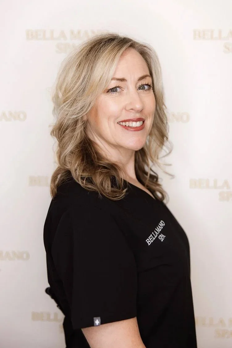 Woman with blonde hair smiling, wearing a black spa uniform with 'BELLA A MANO SPA' embroidered, standing in a spa reception area with a chandelier overhead and pink flowers in the background.