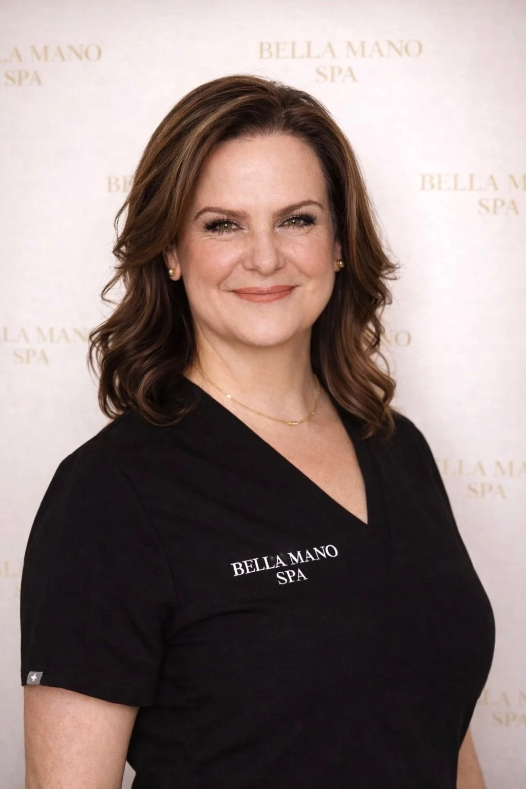 A woman with brown, wavy hair in a beige blazer and jewelry stands at a white marble reception desk with a bouquet of pink and green flowers. She's smiling in a well-lit, elegant interior.