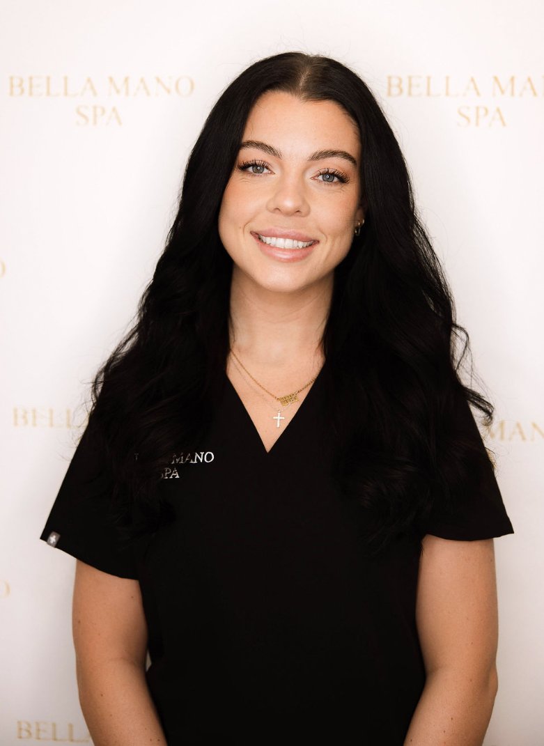 A woman with long black hair and a black uniform standing behind a reception desk, smiling at the camera, with flowers and a chandelier in the background.