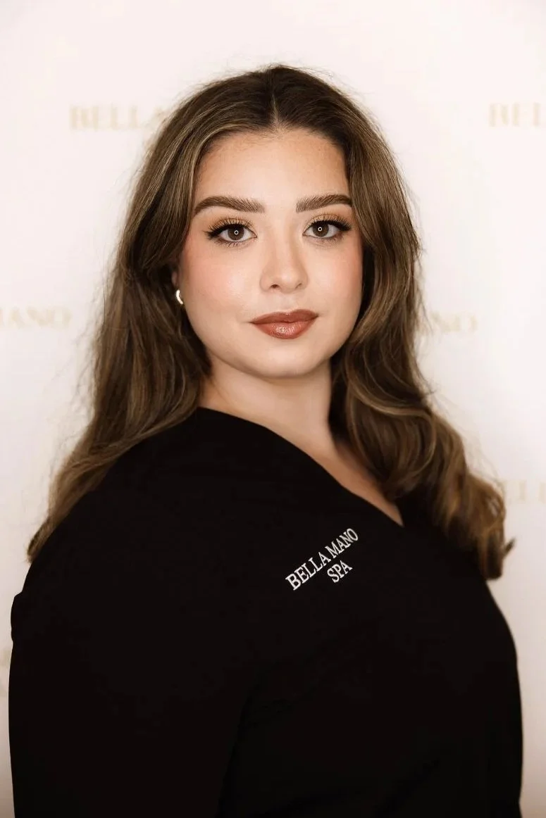 A woman with long brown hair and makeup, wearing a black scrub top that says "Bella Mano Spa," standing in a spa reception area with a chandelier above her.