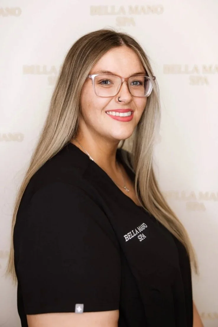 A woman with long light brown hair, glasses, and light skin, wearing a black uniform with "BELLA MANO SPA" embroidery, standing indoors in front of a large crystal chandelier, smiling at the camera.