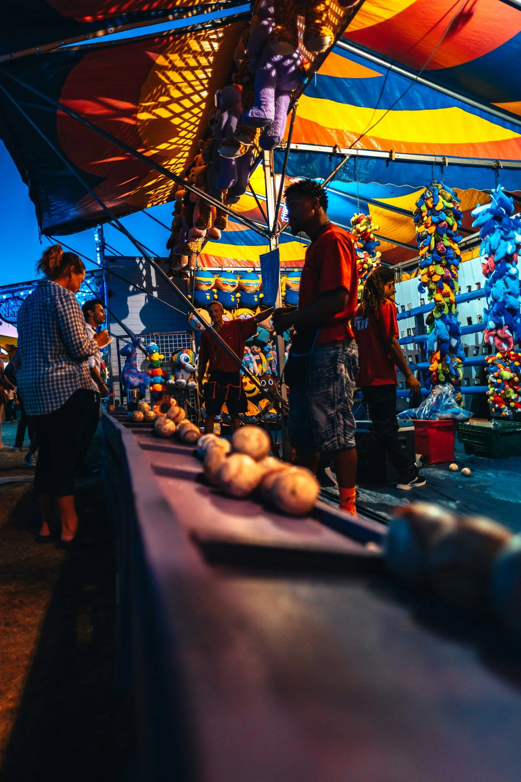 People at a carnival game booth with stuffed animal prizes and colorful decorations, under a vibrant striped canopy at dusk.