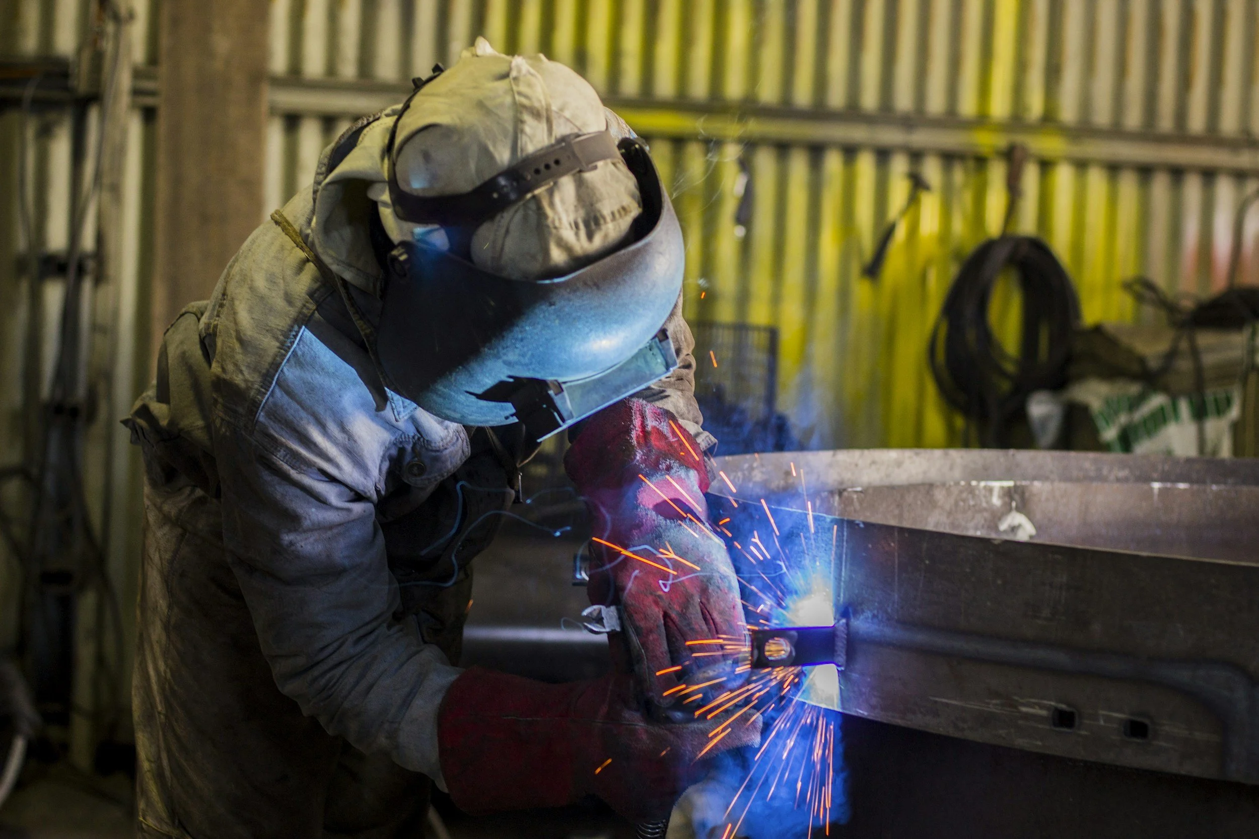 A welder wearing a helmet and protective gloves welding a metal structure inside a workshop.