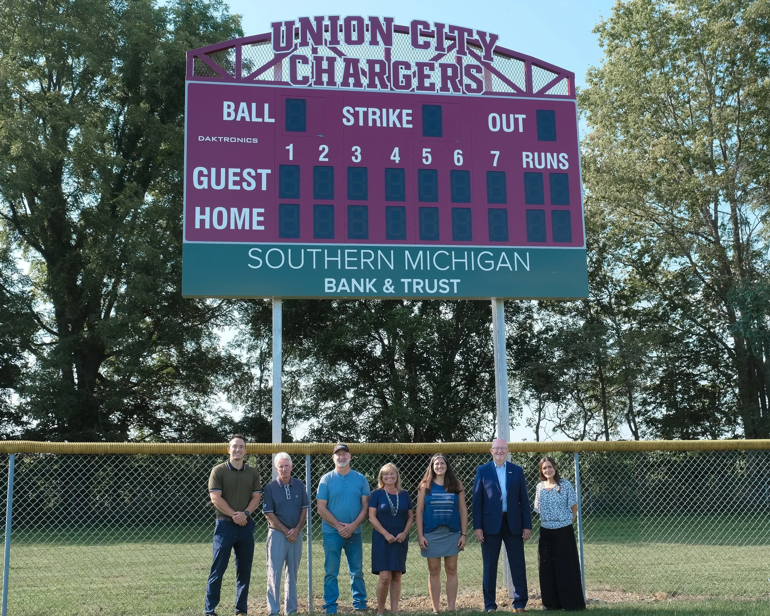 Group of six people standing in front of a large purple sports scoreboard at a baseball field, with trees in the background.